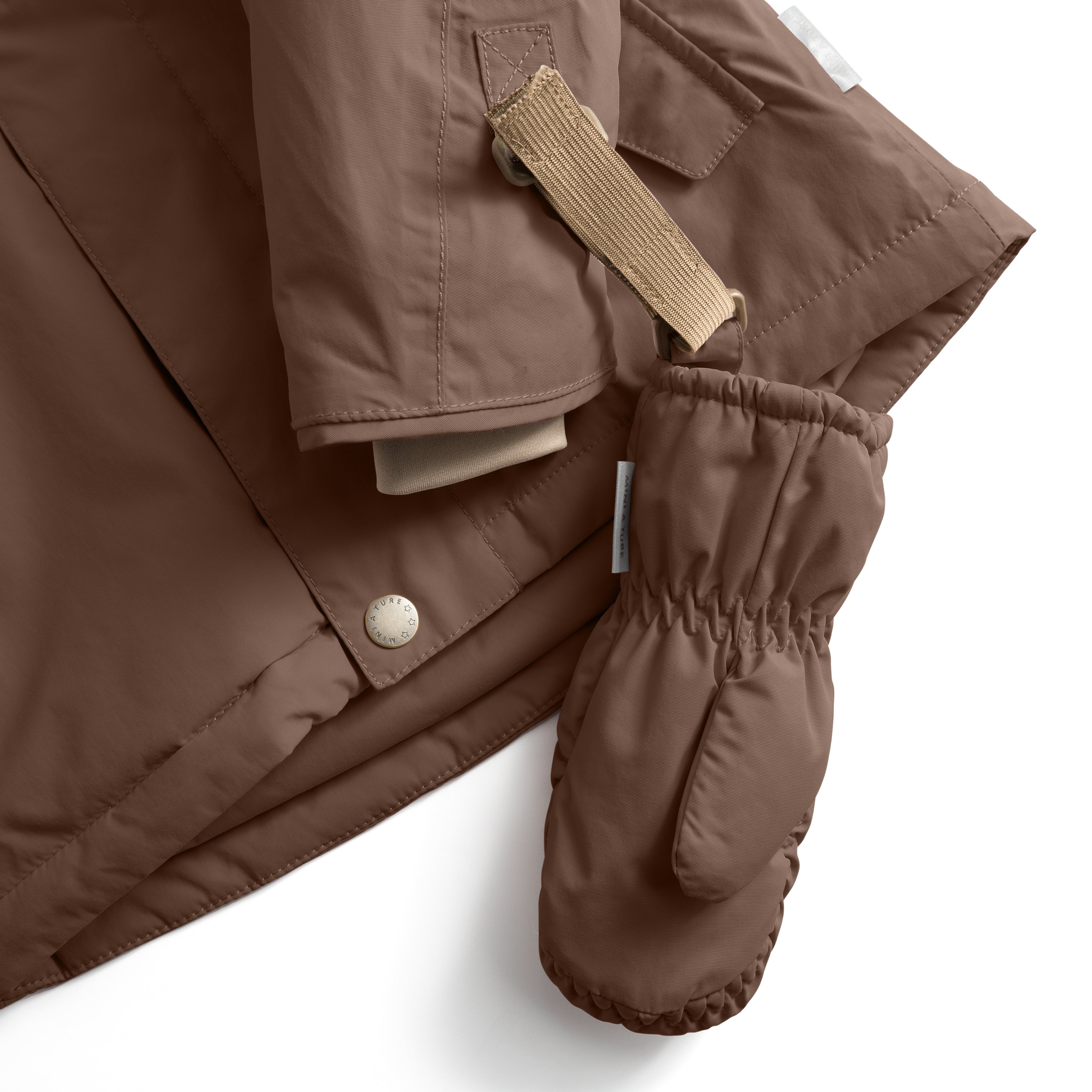 MATCORDT fleece lined mittens in warm brown shown attached to a matching jacket sleeve via the beige velcro strap threaded through the triangular D-loop tab, with the elastic gathered cuff visible at the wrist opening and a press-stud jacket closure alongside.
