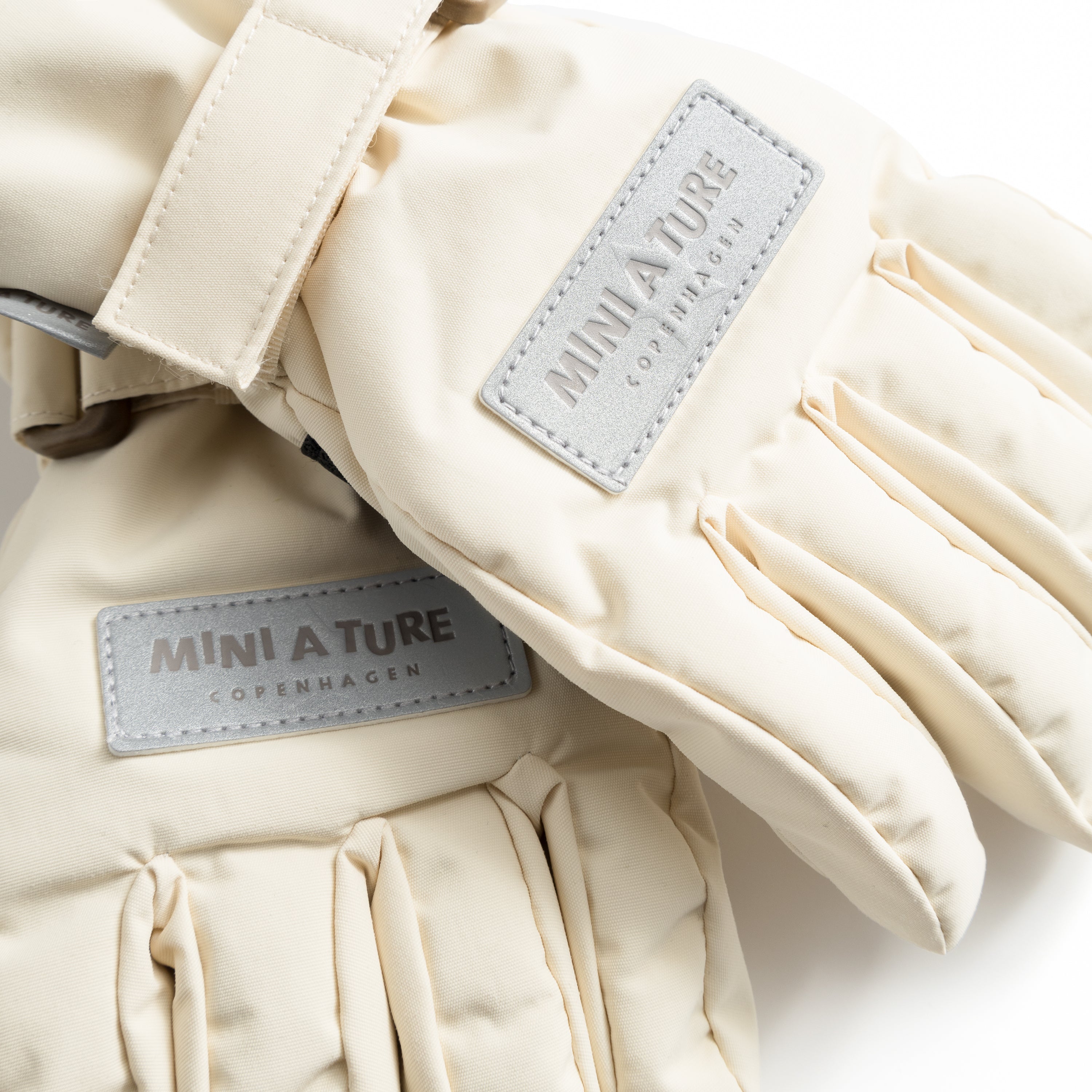 MATCELIO gloves in cream with embossed MINI A TURE Copenhagen labels on the extended cuff, photographed flat to show the waterproof recycled nylon outer shell, THERMOLITE padding construction and adjustable wrist strap with buckle closure.