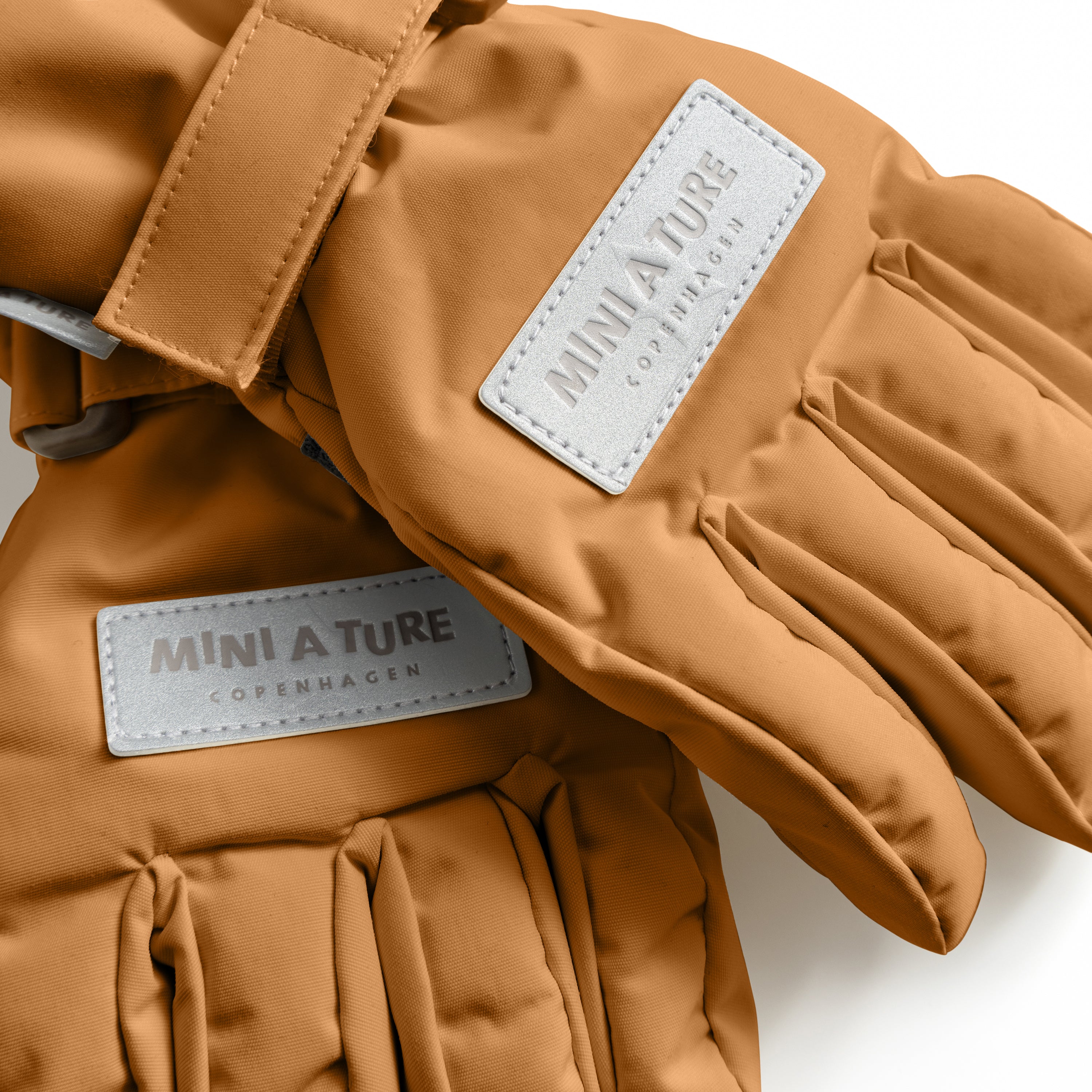 MATCELIO gloves in caramel brown with embossed MINI A TURE Copenhagen labels on the back of each hand and extended cuff, photographed flat to show the waterproof recycled nylon shell and THERMOLITE padding construction.