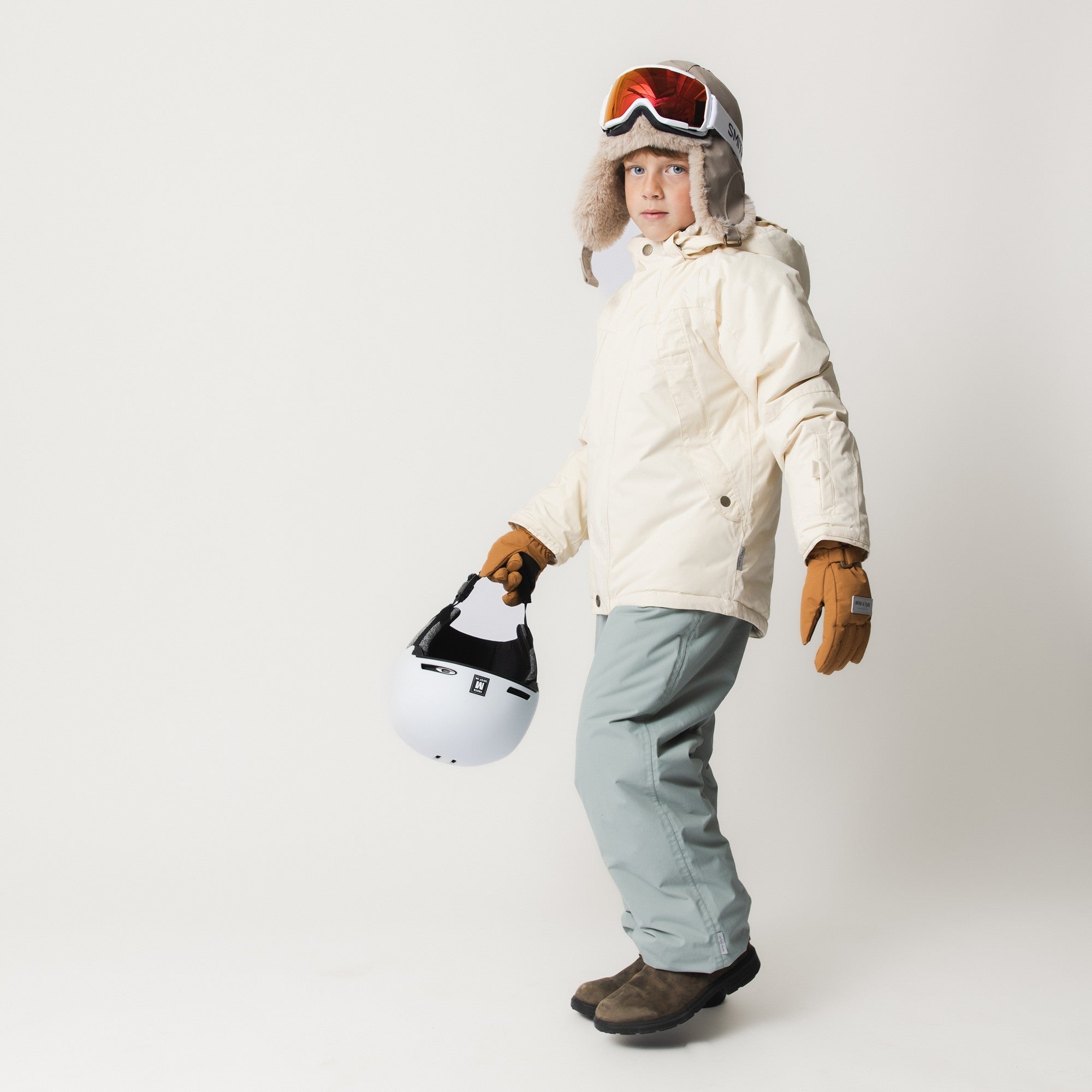 MATCELIO gloves in caramel brown worn with a cream ski jacket and pale blue trousers, showing the extended cuff covering the wrist and the glove's fit during active winter use.