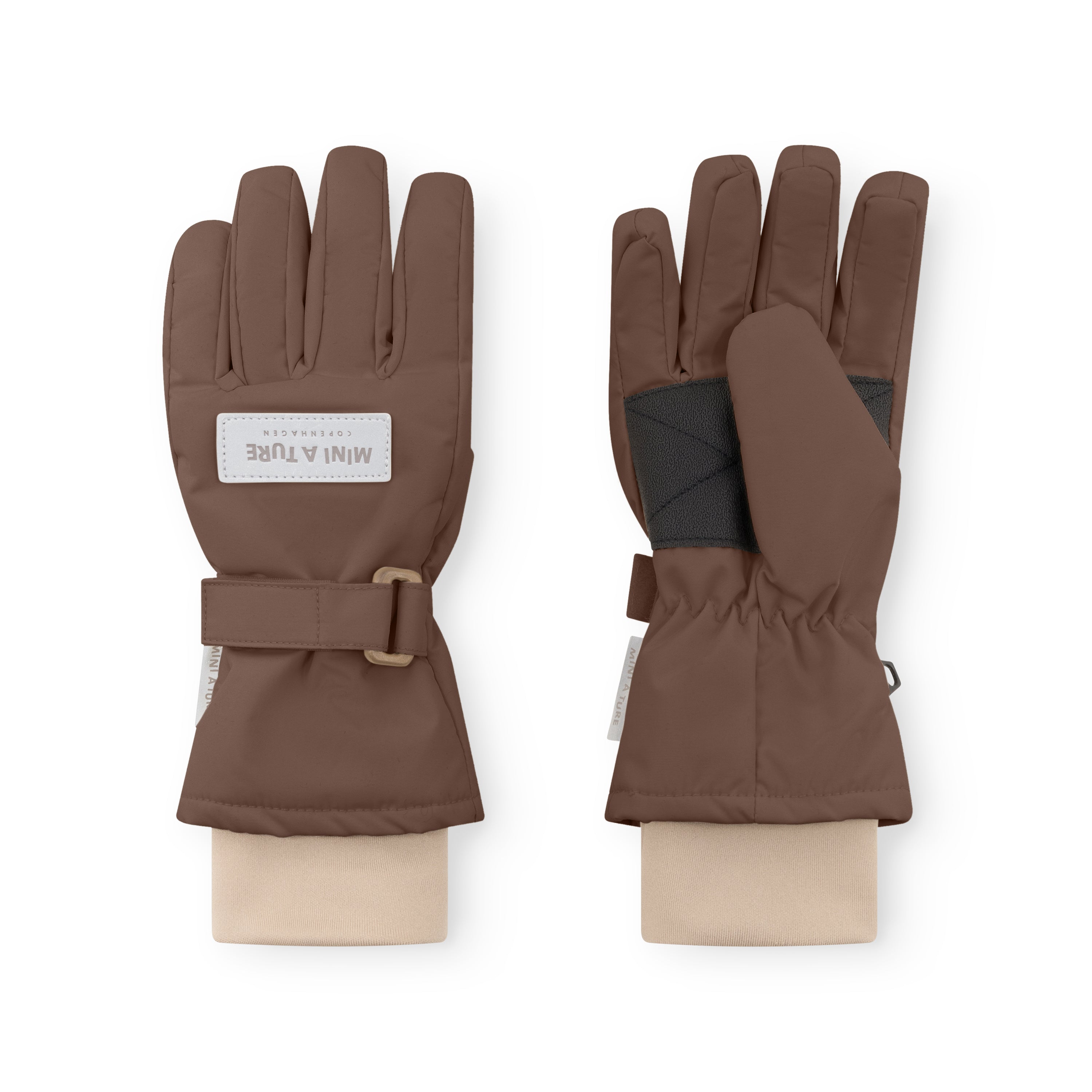 MATCELIO gloves in warm brown displayed on neutral fabric wrists, showing both palm and back views with the white embossed MINI A TURE Copenhagen label on the back of hand, black elasticated internal wrist cuff visible beneath the extended cuff, and the adjustable buckle strap fastened on the outer cuff.