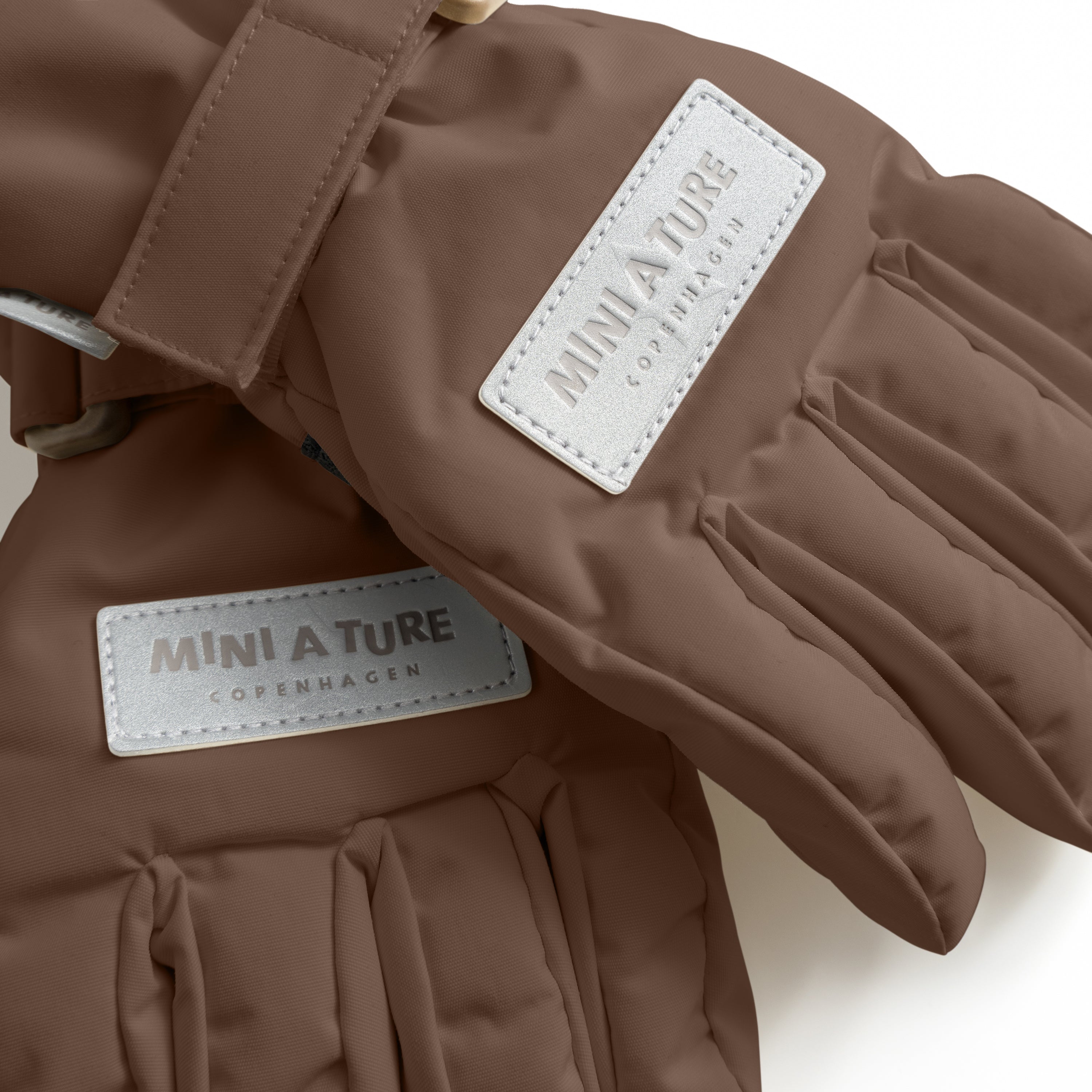 MATCELIO gloves in warm brown with white embossed MINI A TURE Copenhagen labels on both the back of hand and extended cuff, photographed flat to show the waterproof recycled nylon outer shell, THERMOLITE padding construction and adjustable wrist strap with buckle closure.
