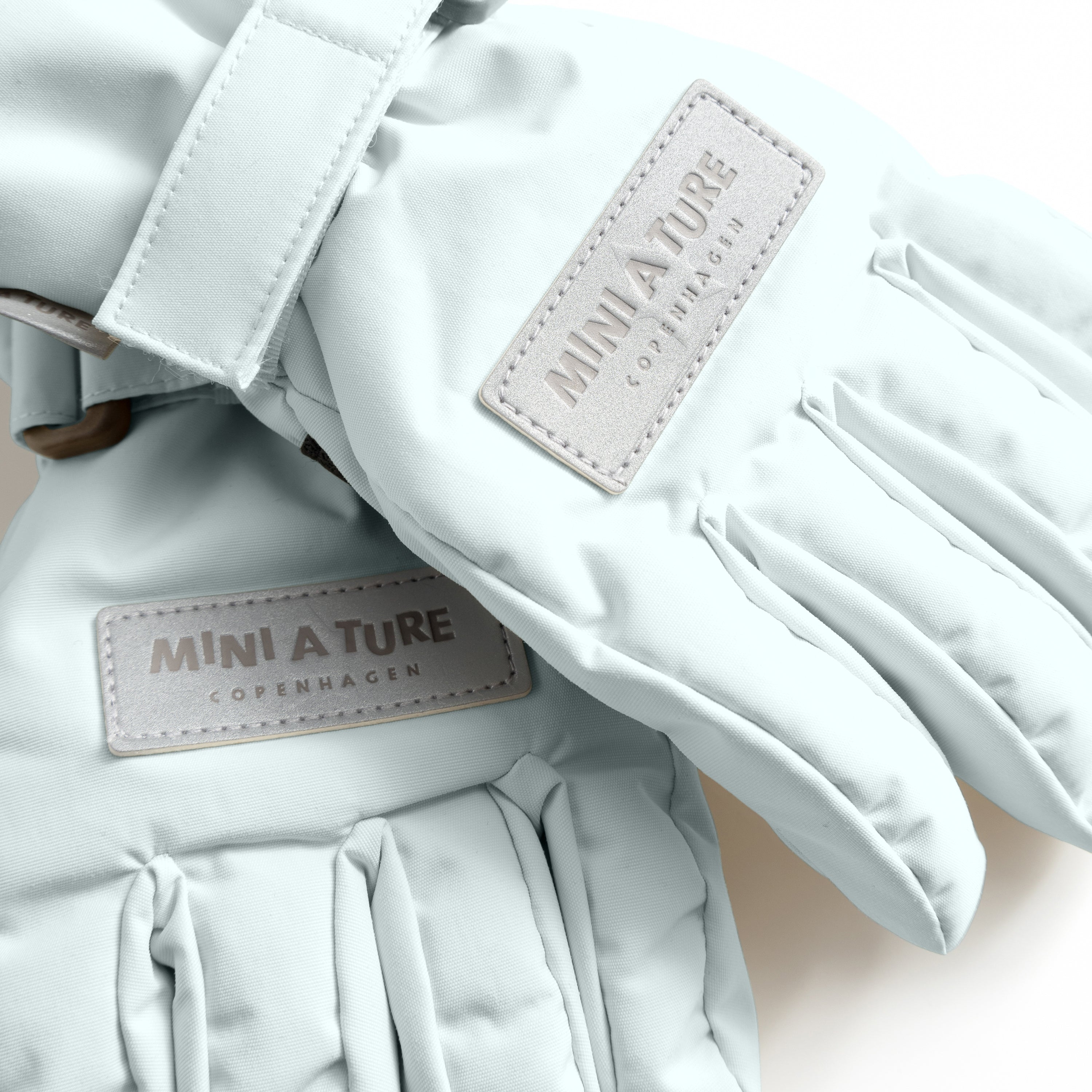 MATCELIO gloves in pale blue-grey photographed flat to show the waterproof recycled nylon outer shell, THERMOLITE padding construction and white embossed MINI A TURE Copenhagen labels on both the back of hand and extended cuff, with the adjustable wrist strap with buckle closure visible.