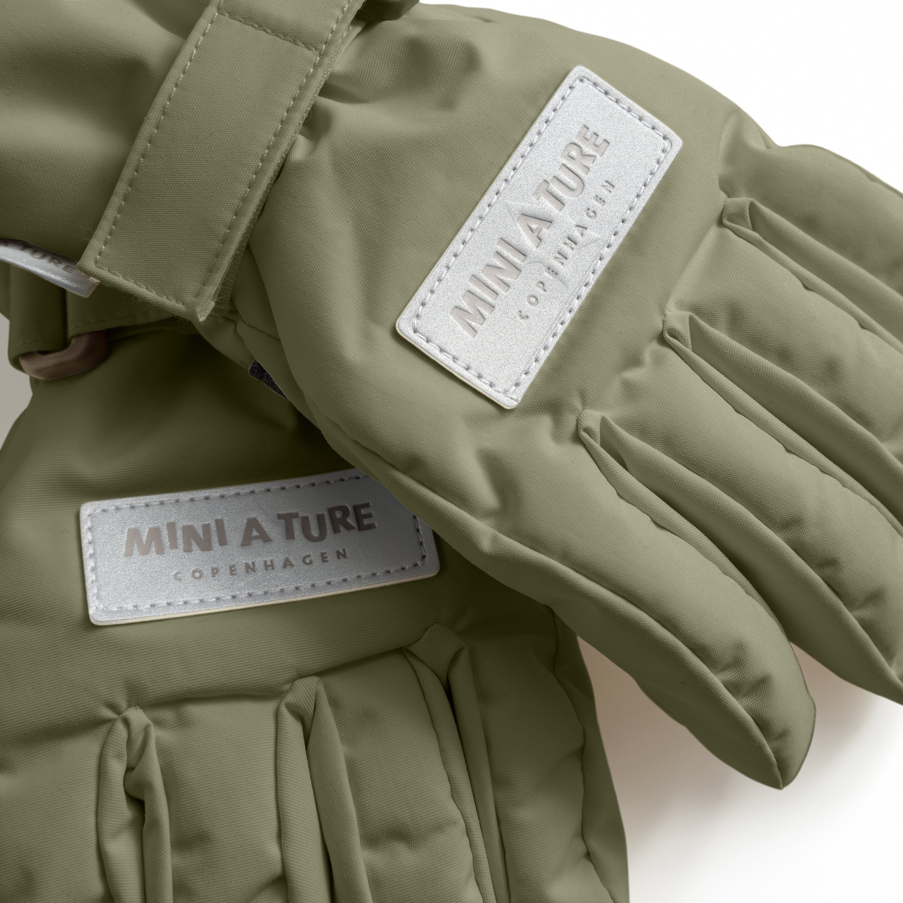 MATCELIO gloves in olive green with white embossed MINI A TURE Copenhagen labels on both the back of hand and extended cuff, photographed flat to show the waterproof recycled nylon outer shell, THERMOLITE padding construction and adjustable wrist strap with buckle closure.
