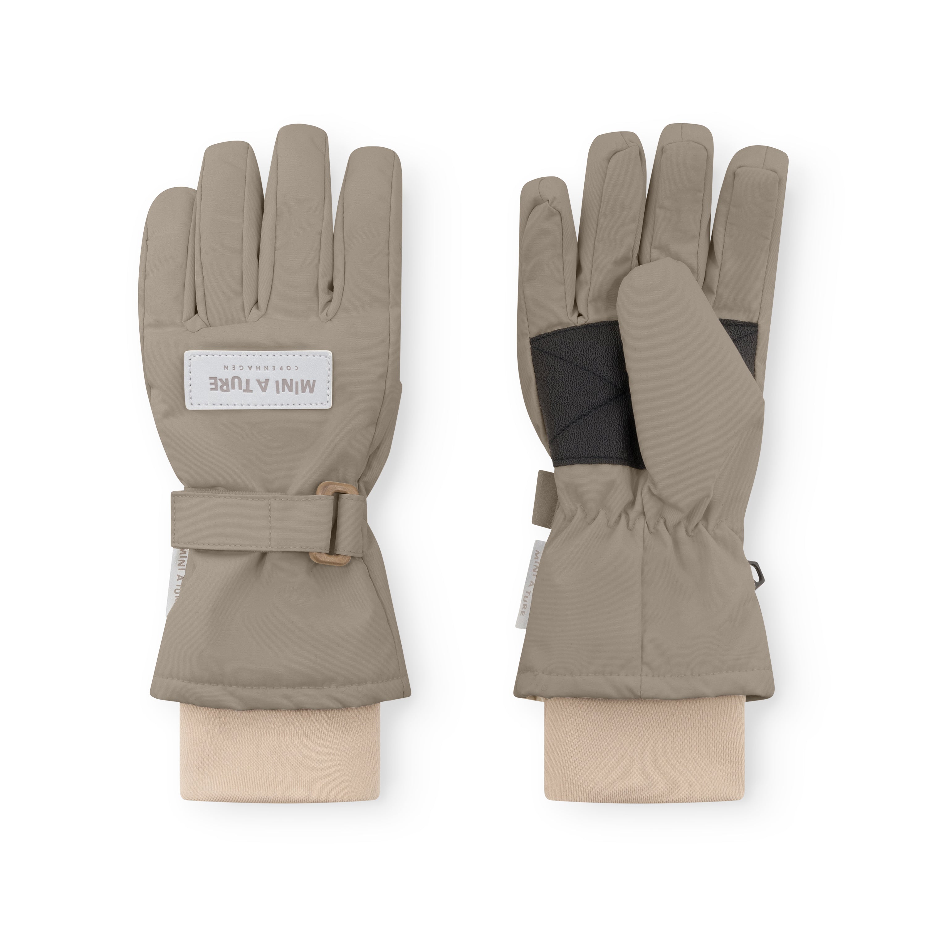 MATCELIO gloves in warm taupe displayed on neutral fabric wrists, showing both palm and back views with the white embossed MINI A TURE Copenhagen label on the back of hand, black elasticated internal wrist cuff visible beneath the extended cuff, and the adjustable buckle strap fastened on the outer cuff.