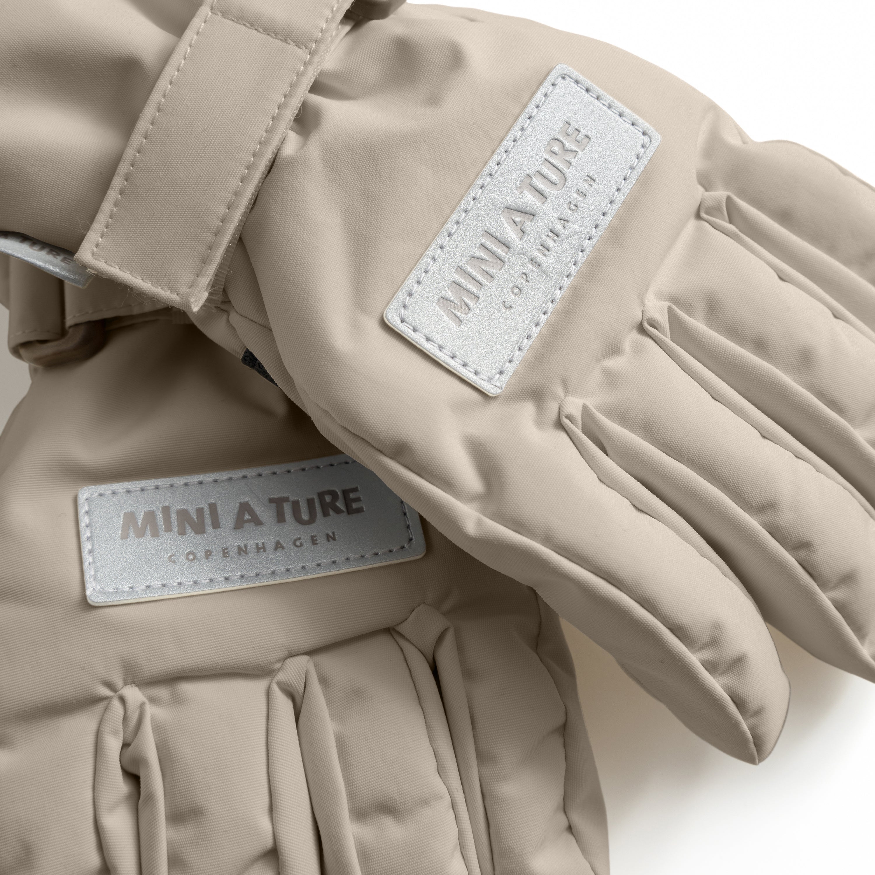 MATCELIO gloves in warm taupe photographed flat to show the waterproof recycled nylon outer shell, THERMOLITE padding construction and white embossed MINI A TURE Copenhagen labels on both the back of hand and extended cuff, with the adjustable wrist strap with buckle closure visible.