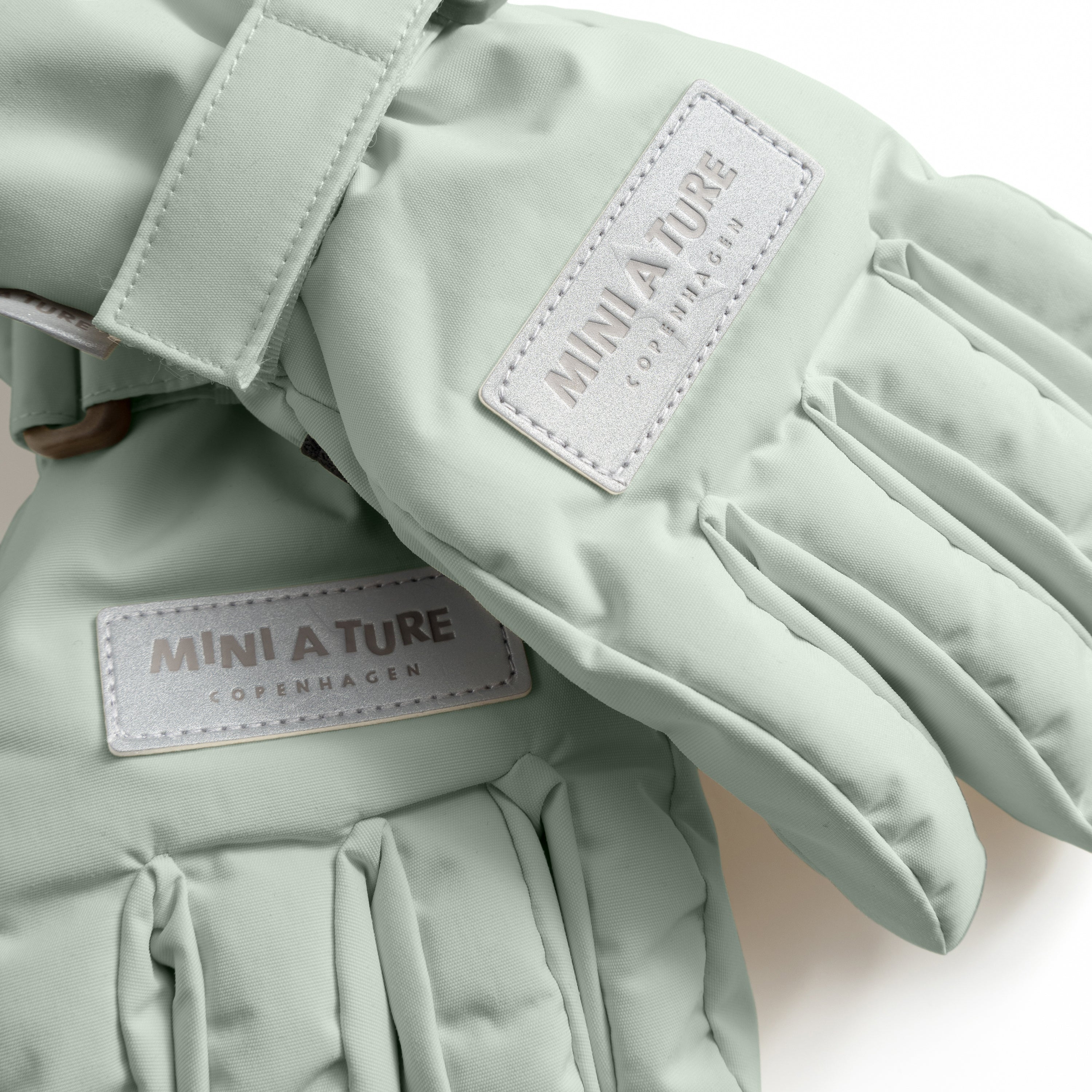 MATCELIO gloves in soft sage green with embossed MINI A TURE Copenhagen labels on the extended cuff, photographed flat to show the waterproof recycled nylon outer shell, THERMOLITE padding construction and adjustable wrist strap with buckle closure.