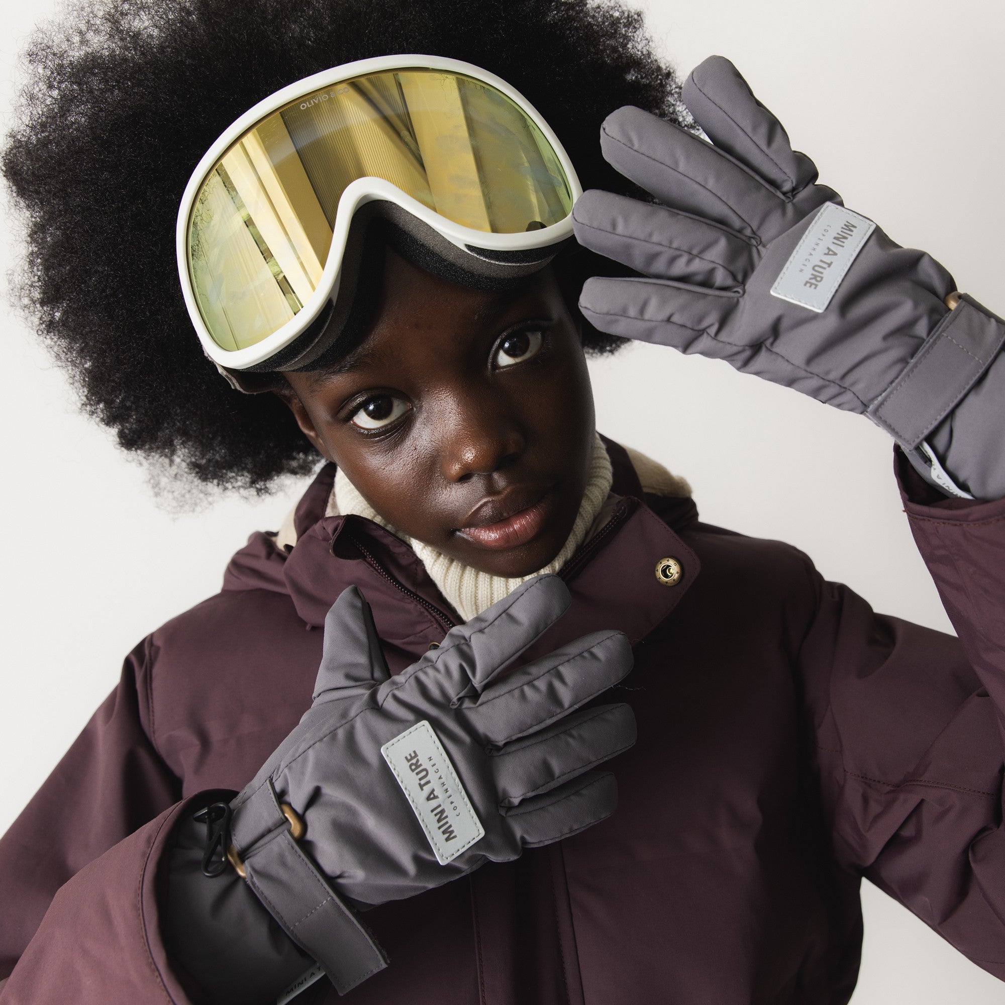 MATCELIO gloves in grey with adjustable wrist strap and reflective MINI A TURE logo, shown worn with ski goggles and winter jacket.