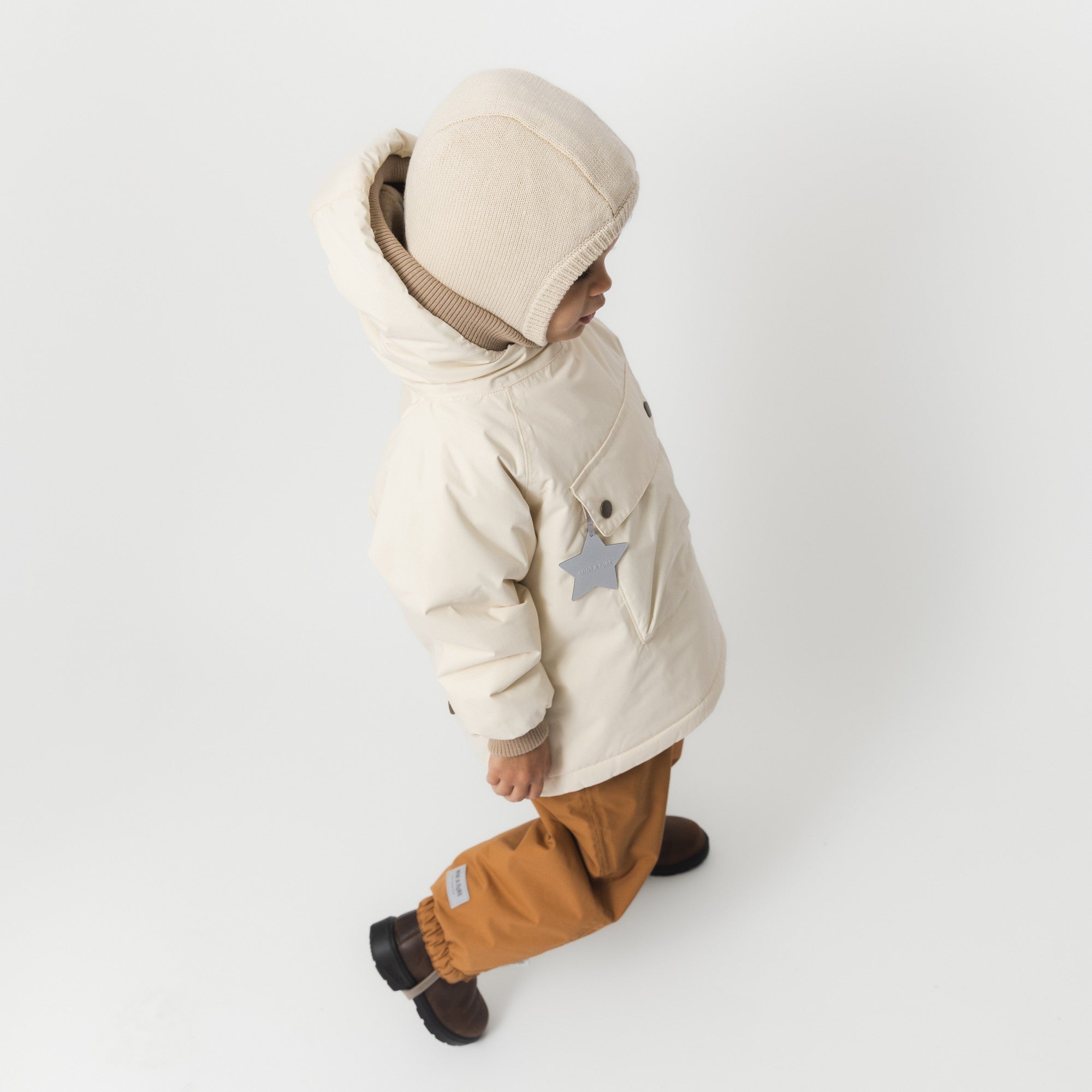 MATBABYWEN anorak in cream shown from the side on a toddler, displaying the ribbed hood and cuff detail, snap-button front pocket with reflective star tag, and the coat's length over mustard trousers.
