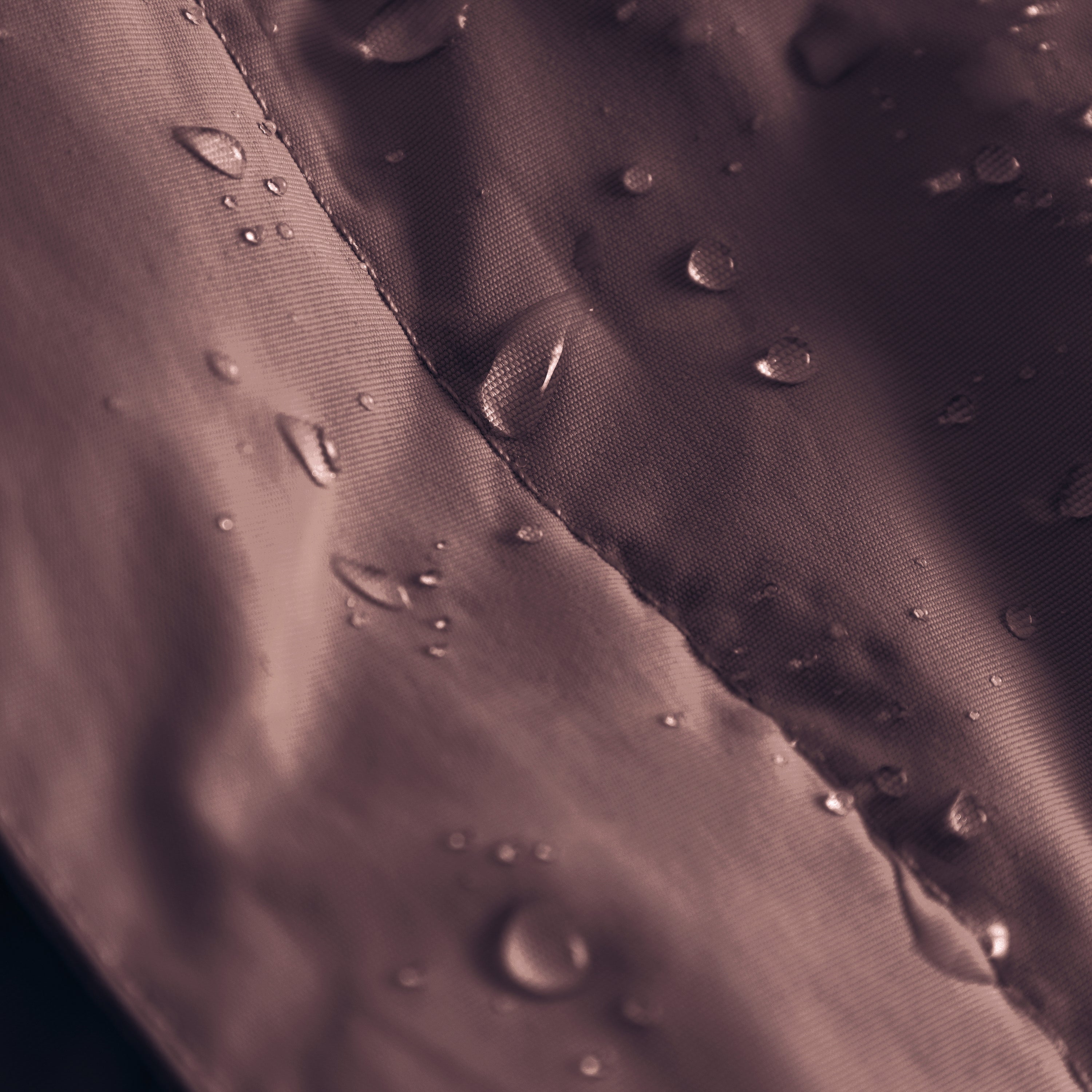 Close-up of the MATBABYWEN anorak in dark plum showing water droplets beading on the recycled polyamide outer shell treated with fluorine-free Bionic Finish Eco, with a detailed view of the taped seam running diagonally across the waterproof fabric.