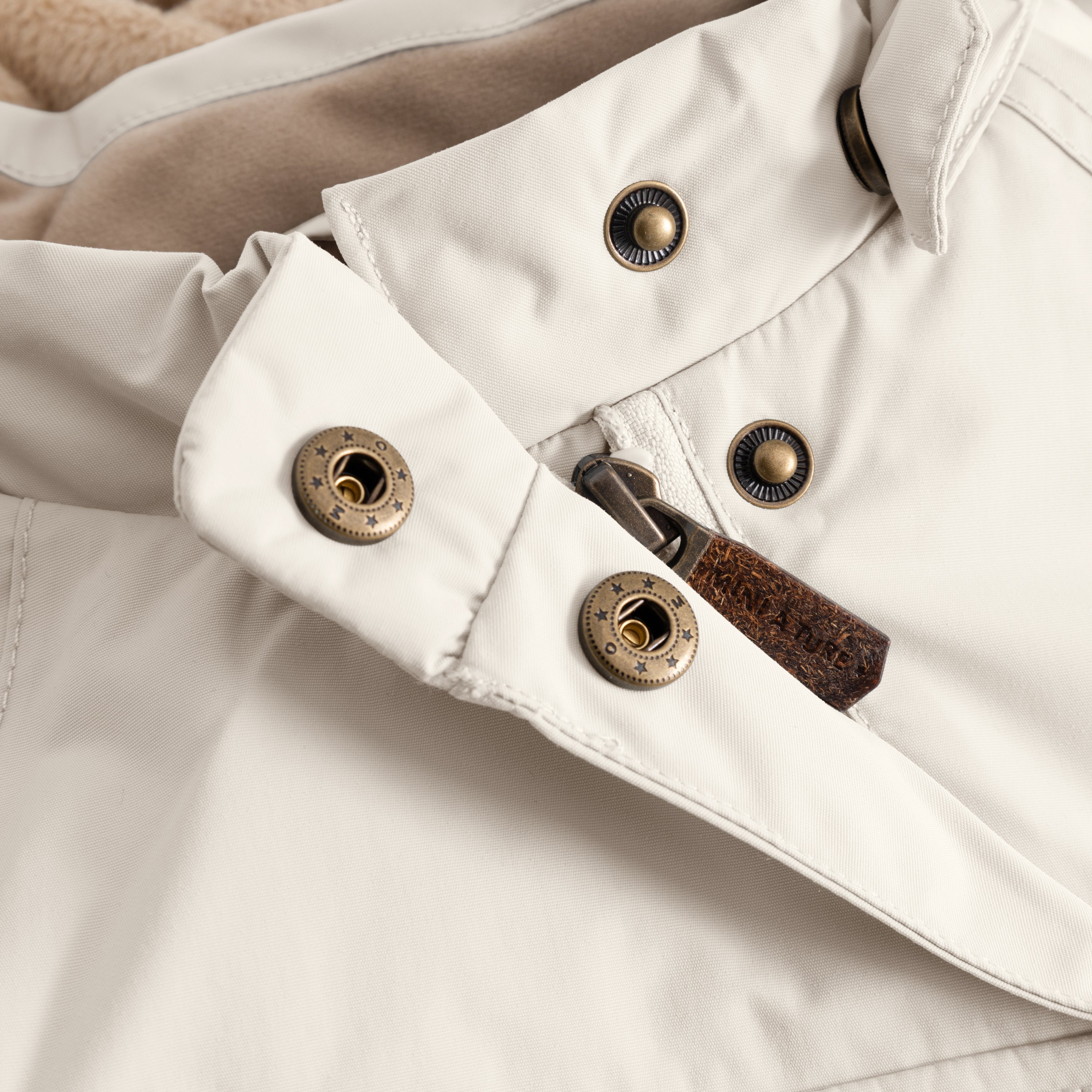 Detail of the MATWANG fleece-lined winter jacket's right sleeve cuff showing the bronze press-stud fastening system with radiating line detail, brown leather zip pull, and the adjustable snow cuff with thumb opening visible at the wrist, all in soft cream recycled nylon.
