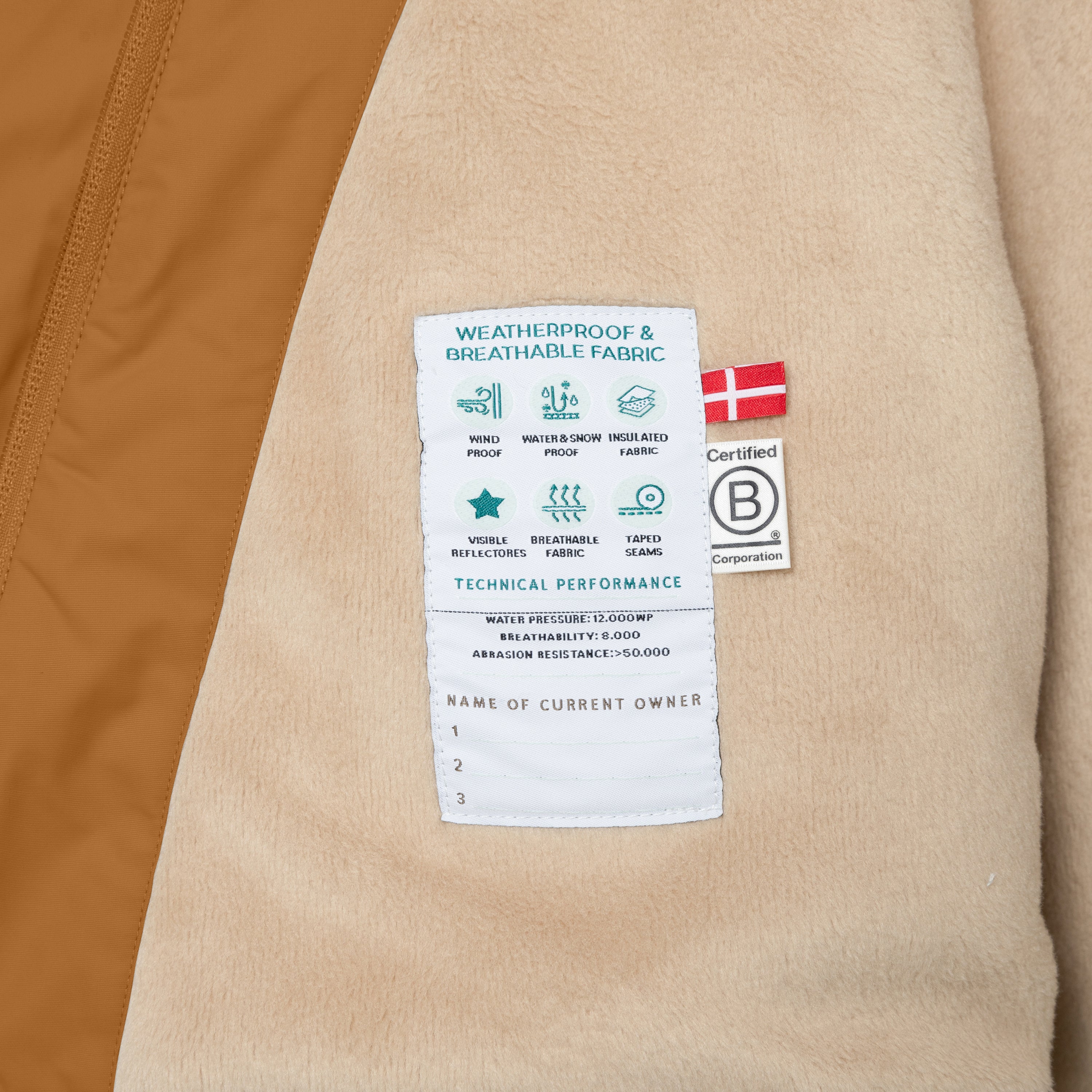 Interior label of the MATWANG fleece-lined winter jacket showing technical performance specifications including 12,000mm water pressure rating, 8,000 g/m²/24h breathability and 50,000+ Martindale abrasion resistance, alongside a Danish flag and B Corp certification logo, with beige fleece lining and warm brown outer shell visible.