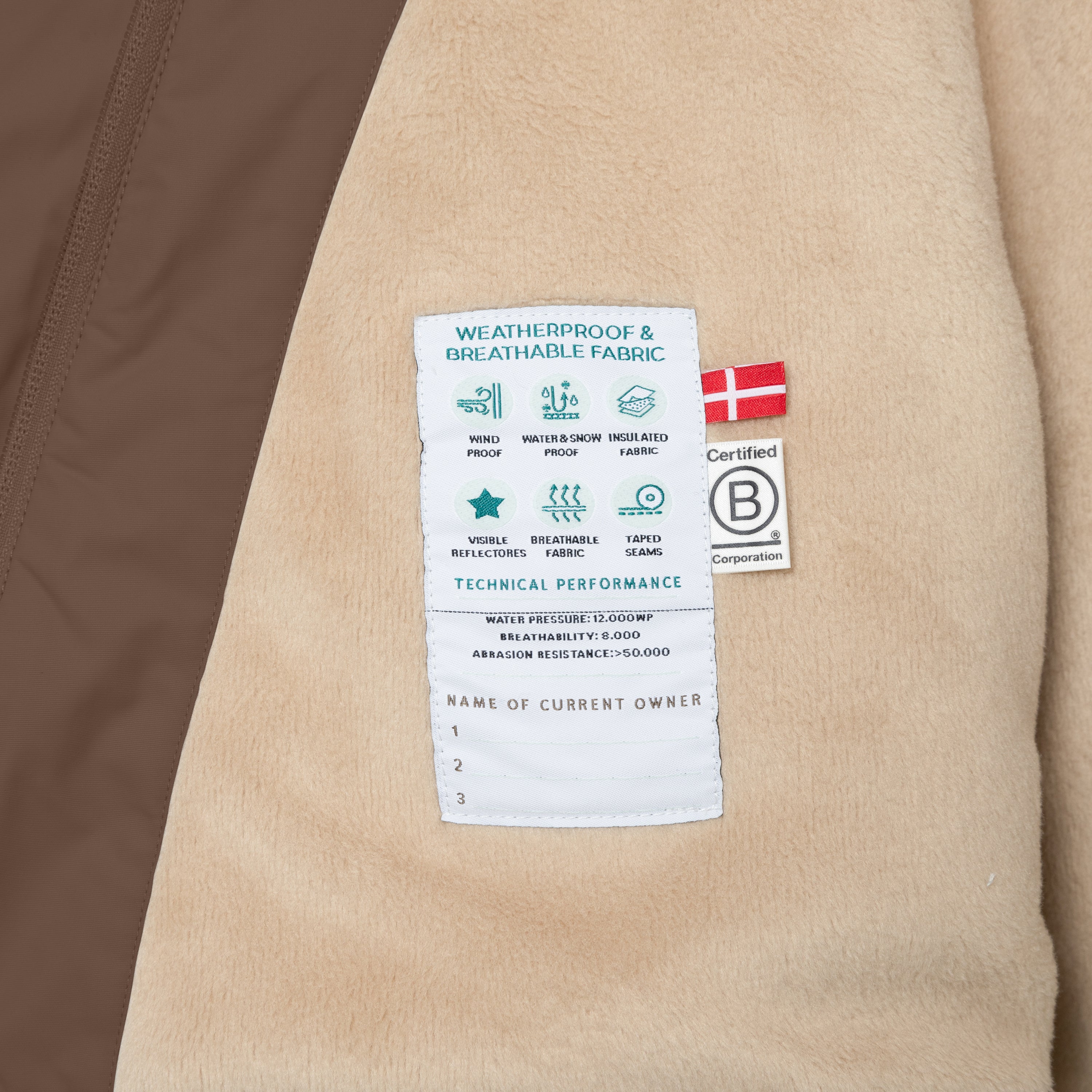 Interior technical label of the MATWANG fleece-lined winter jacket sewn onto the beige fleece lining, displaying weatherproof and breathable fabric icons, windproof and water-resistant symbols, visible reflectors and taped seams indicators, technical specifications including 12,000mm water pressure, 8,000 breathability rating and over 50,000 abrasion resistance, Danish flag, B Corp certification logo, and a section for recording up to three owner names.