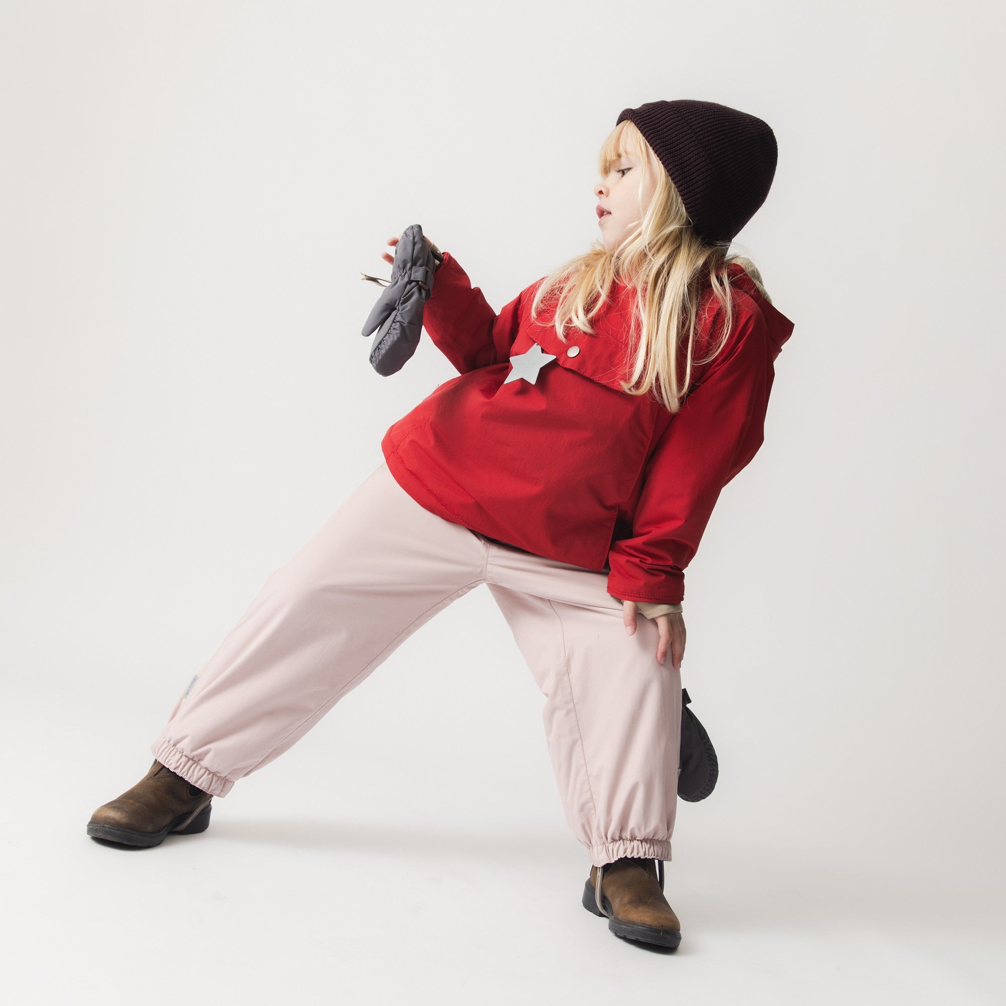 Child wearing the MATWANG fleece-lined winter jacket in red paired with pale pink trousers and a dark knitted beanie, crouching whilst holding grey mittens, demonstrating the jacket's freedom of movement and everyday styling versatility.