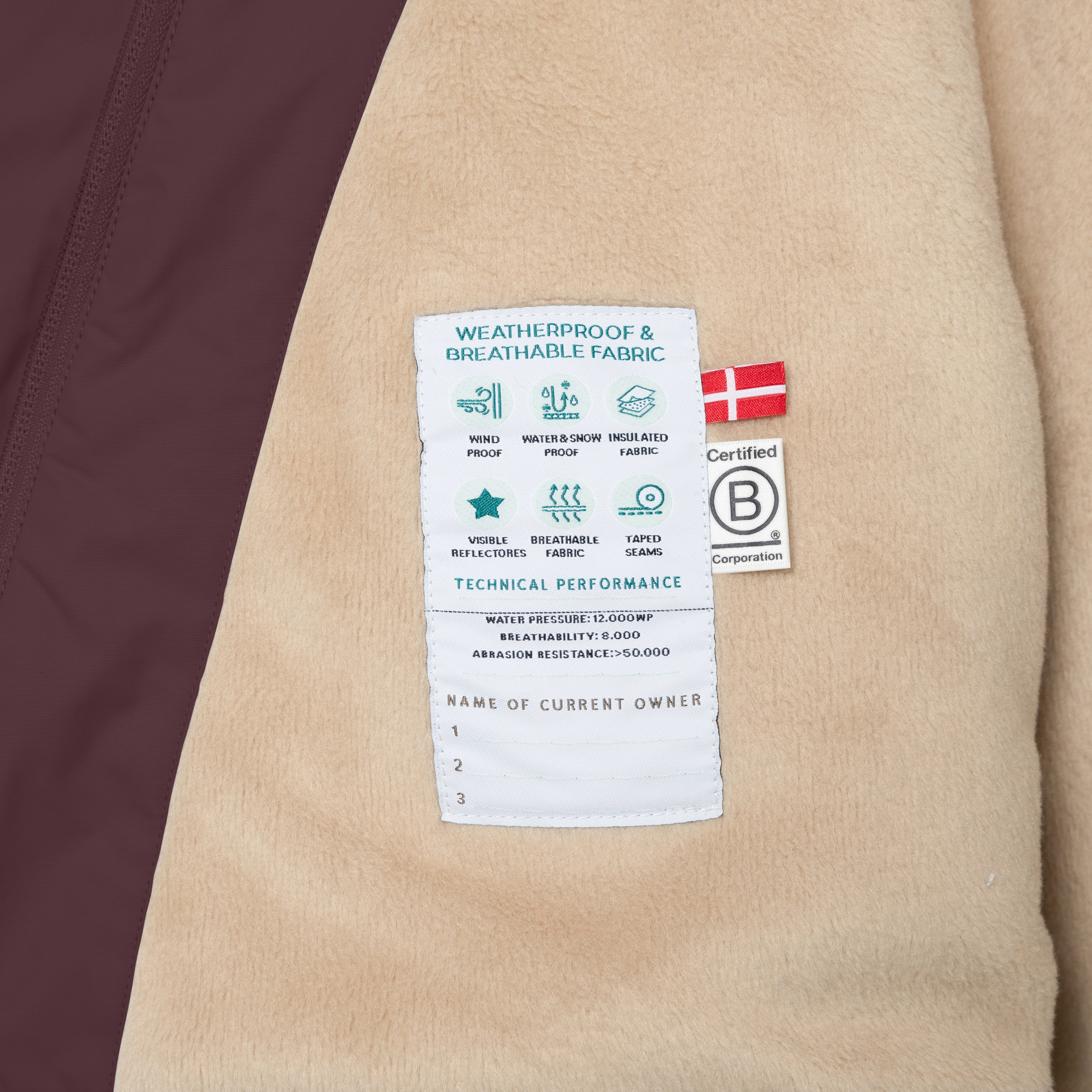 Interior care label of the MATWANG fleece-lined winter jacket sewn onto the beige fleece lining, showing weatherproof and breathable fabric icons, technical specifications including 12,000mm water pressure and 8,000 breathability rating, 50,000+ abrasion resistance, Danish flag, B Corp certification mark, and a section for recording up to three owner names.