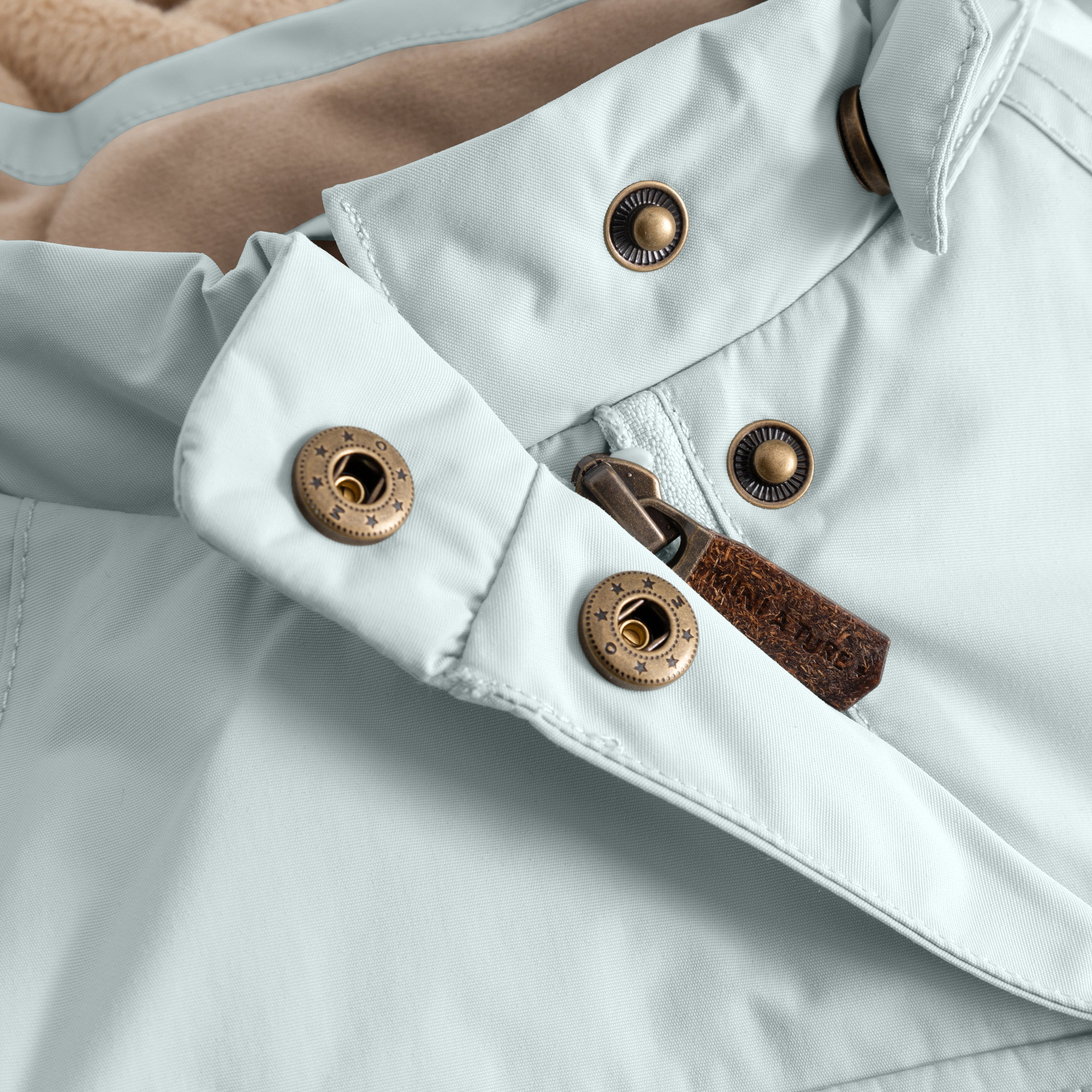 Close-up of the MATWANG fleece-lined winter jacket's adjustable internal waist showing the bronze-toned metal press-stud fastening system with elastic adjustment mechanism and brown fabric pull tab, demonstrating the fit refinement feature on the pale blue waterproof recycled nylon shell.
