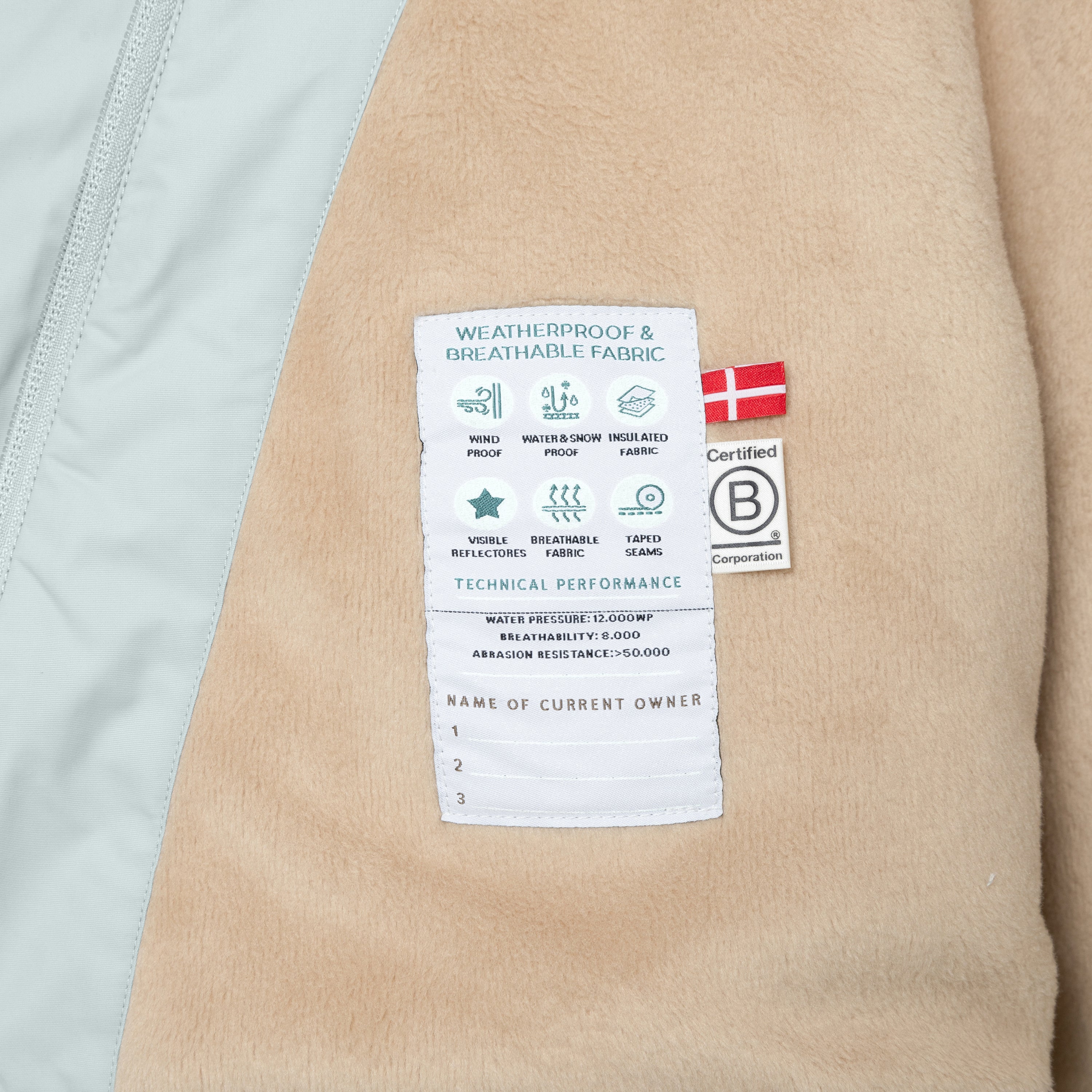 Interior view of the MATWANG fleece-lined winter jacket showing the white technical performance label sewn onto the beige fleece lining, displaying windproof, waterproof and breathability specifications alongside the Danish flag, B Corp certification mark, and a blank owner registration section.