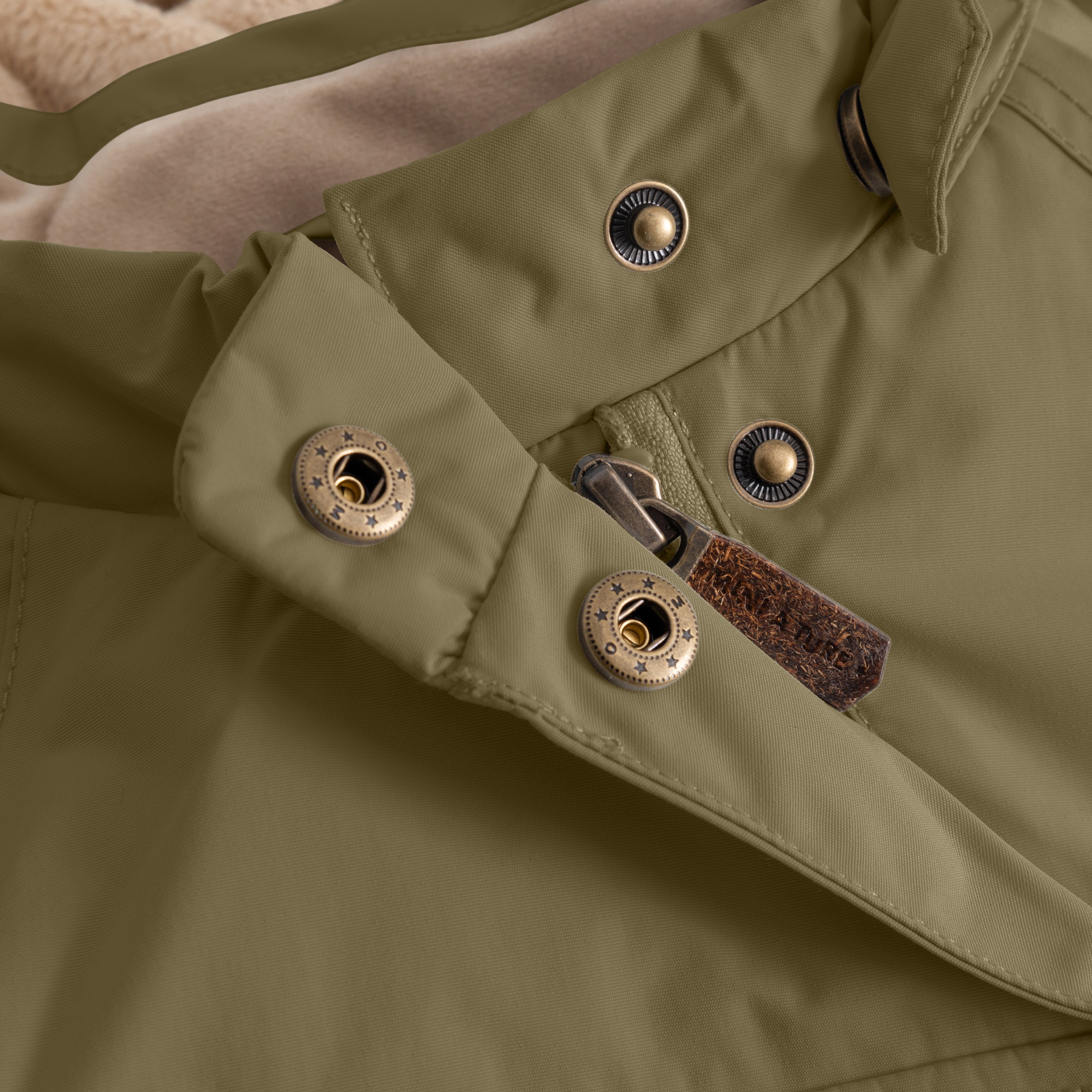 Close-up of the MATWANG fleece-lined winter jacket's detachable hood fastening system showing bronze press studs with radiating line detail, a bronze zip pull with leather tab, and beige fleece lining visible at the hood edge.
