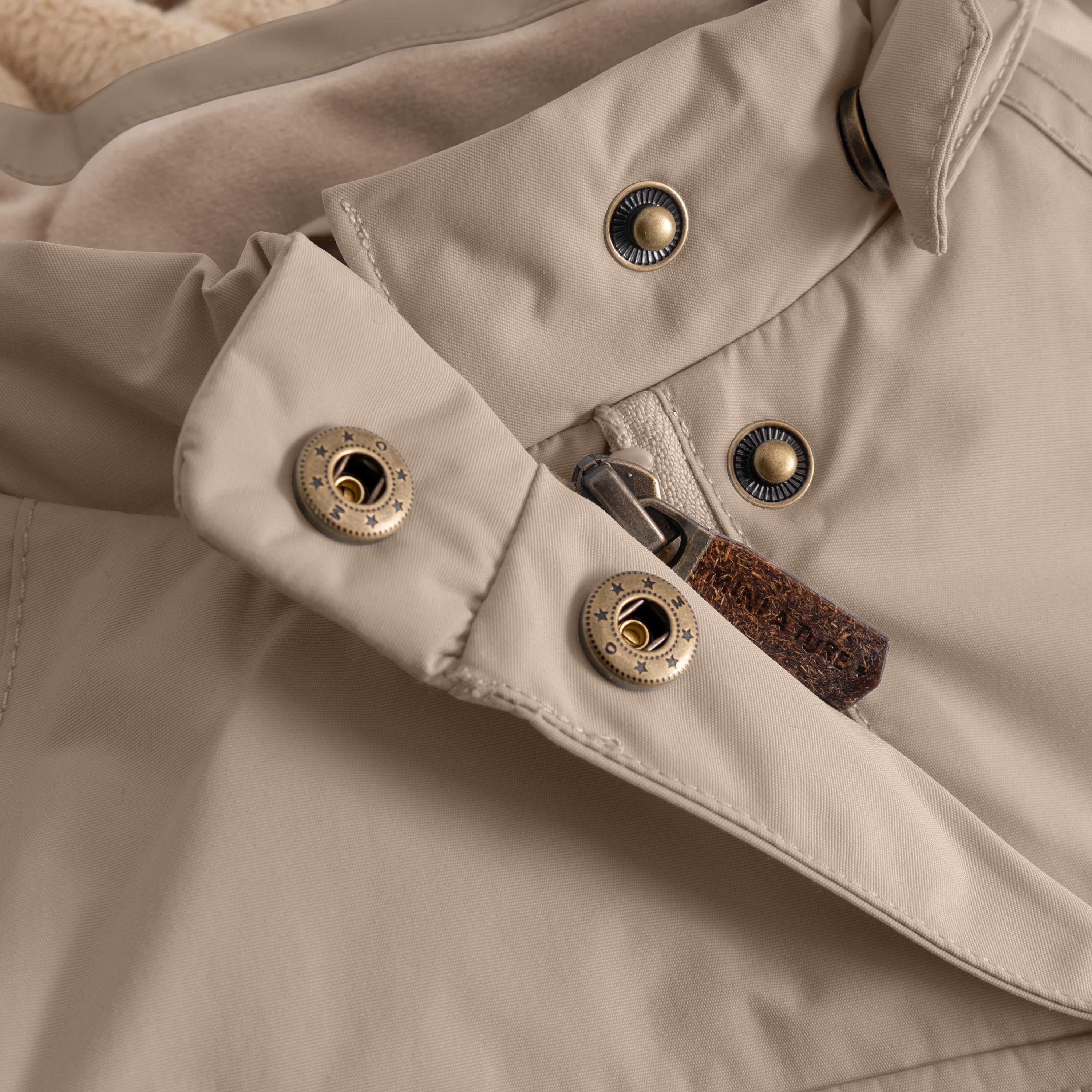 Close-up of a children's winter jacket in neutral beige showing a hidden slanted zip fastening with metal press studs and an adjustable cuff with snap closure, highlighting durable construction details.