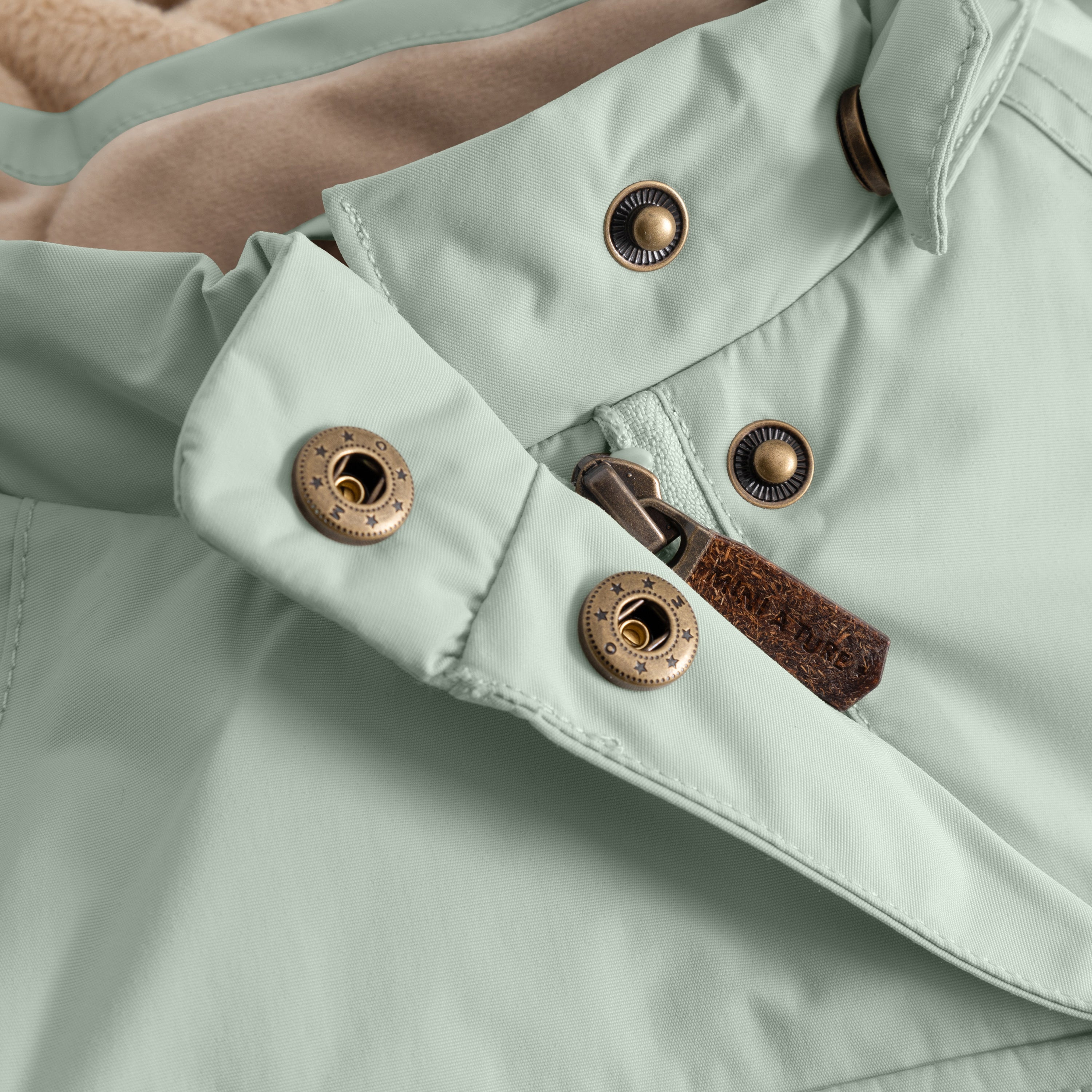 Close-up of the MATWANG fleece-lined winter jacket's left sleeve cuff showing two bronze press studs with radiating line detail, a bronze zip pull with brown leather tab, and the adjustable snow cuff fastening system with beige fleece lining visible at the wrist opening.