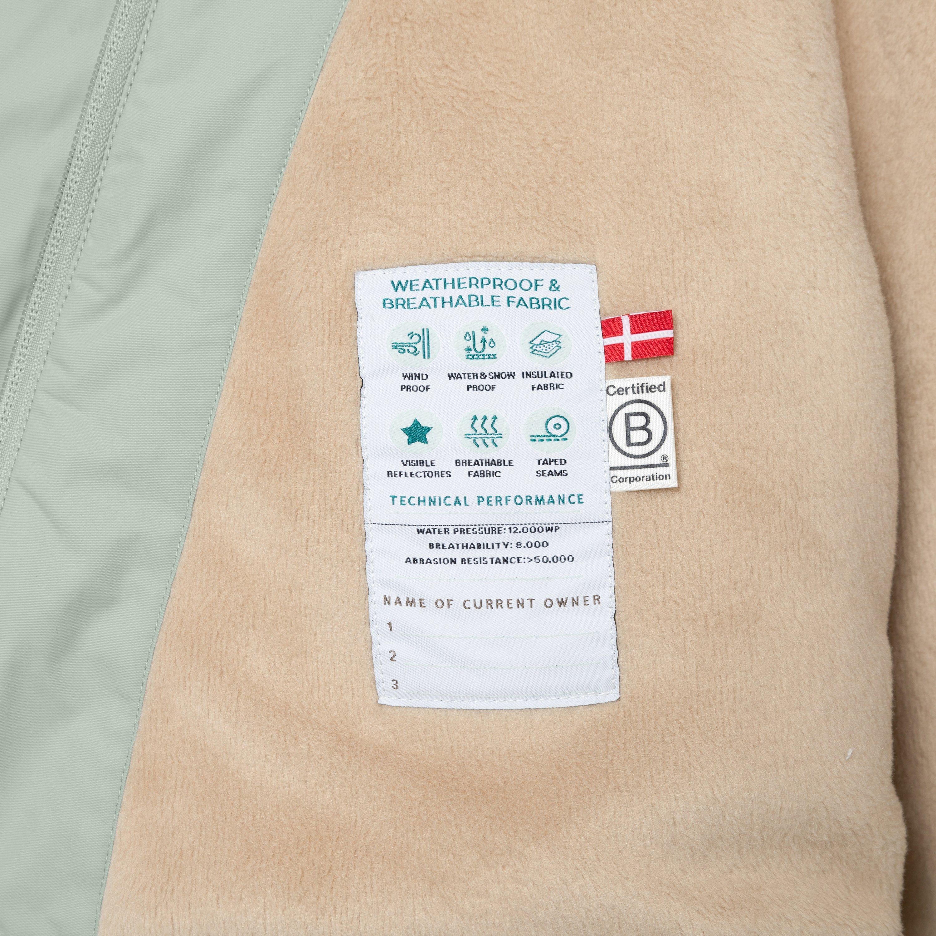 Interior label of the MATWANG fleece-lined winter jacket displaying weatherproof and breathable fabric icons, technical performance ratings including 12,000mm water pressure and 8,000 breathability, abrasion resistance above 50,000, B Corp certification logo with Danish flag, and owner registration section, sewn onto beige fleece lining with sage green outer shell visible at the left edge.
