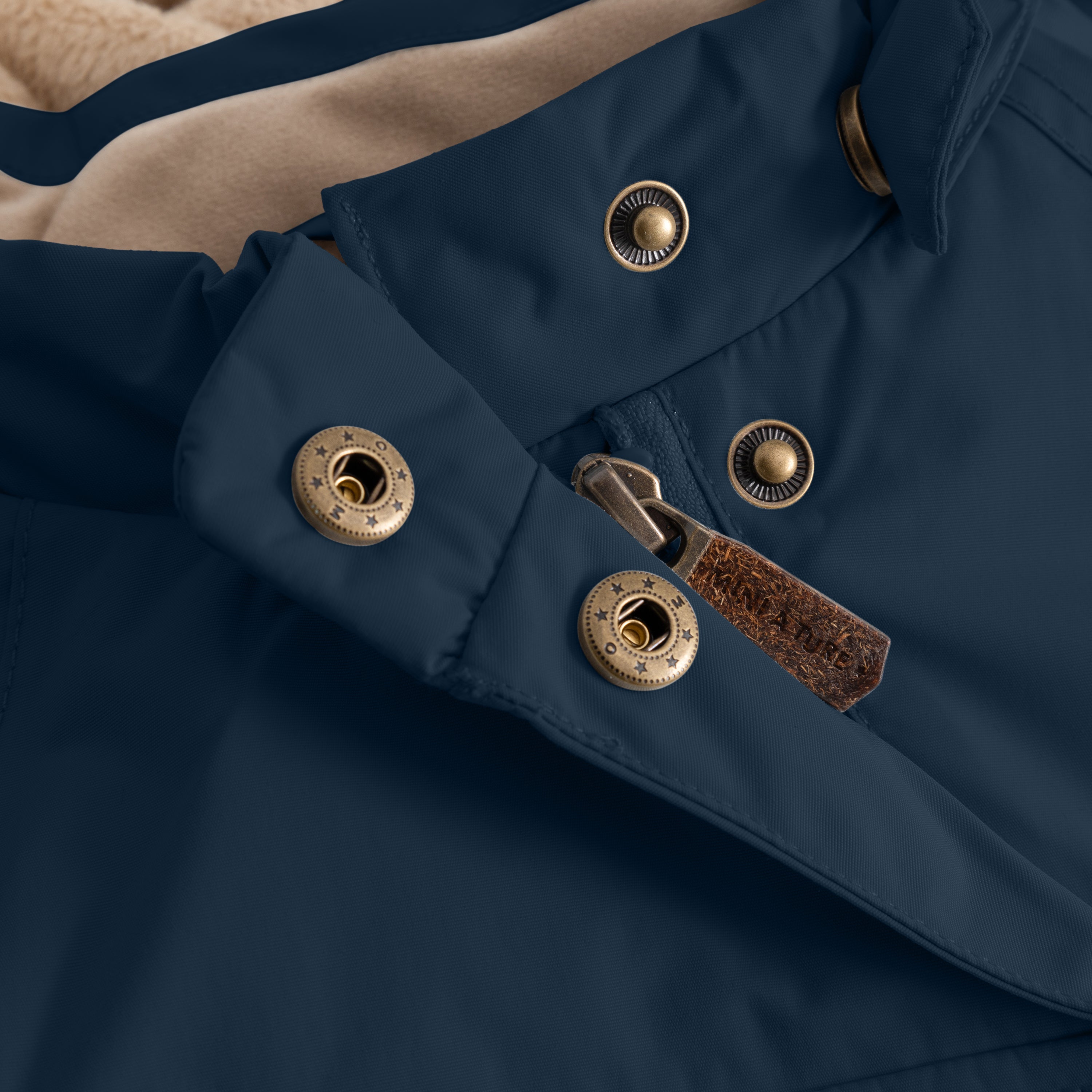 Close-up detail of a dark blue children's winter jacket showing a hidden slanted zip with a leather pull tab and brass press-stud fasteners on a water-repellent recycled nylon shell.