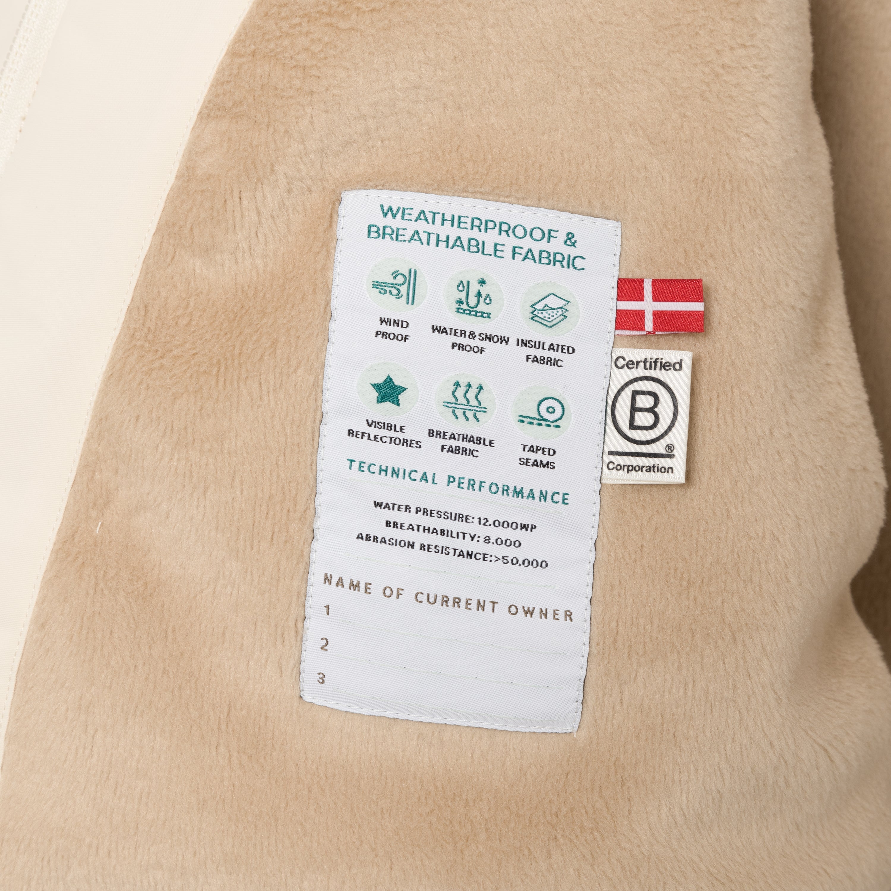 Close-up of the MATWANG fleece lined winter jacket's internal technical label sewn into the beige fleece lining, displaying windproof, waterproof and breathable fabric icons, 12,000mm water pressure rating, 8,000 g/m²/24h breathability, abrasion resistance above 50,000 Martindale, Danish flag, B Corp certification logo, and three numbered lines for recording current owner details.