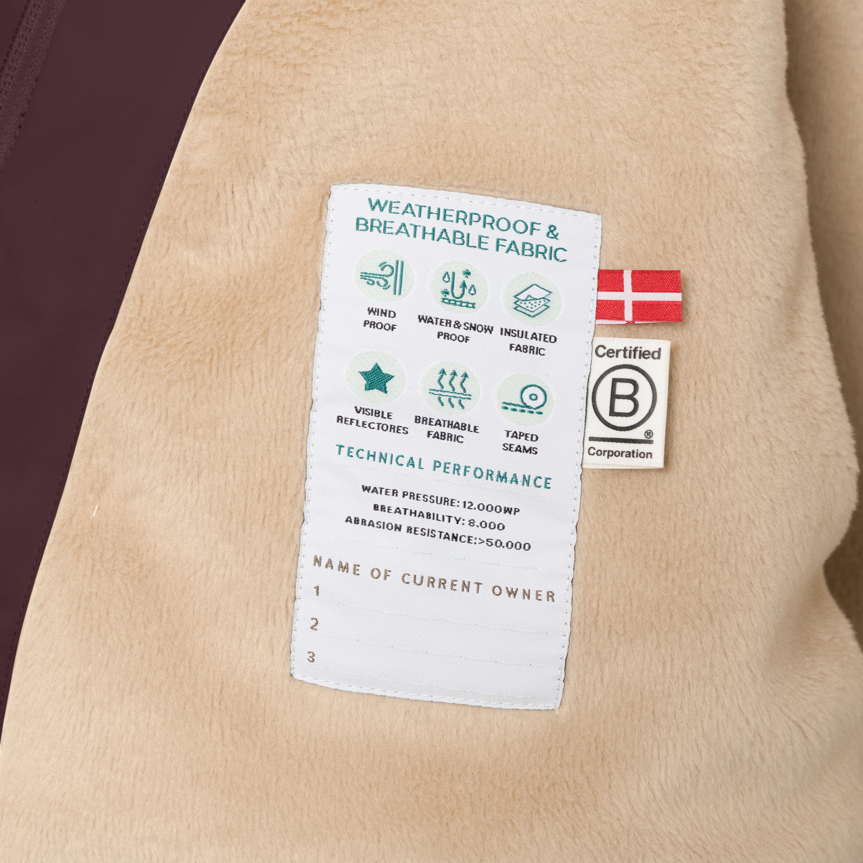 Internal care label of the MATWANG fleece lined winter jacket sewn into the beige fleece lining, showing windproof, waterproof and breathable fabric icons, 12,000mm water pressure rating, 8,000 g/m²/24h breathability, abrasion resistance above 50,000 Martindale, Danish flag, B Corp certification logo, and three numbered spaces for recording current owner details.