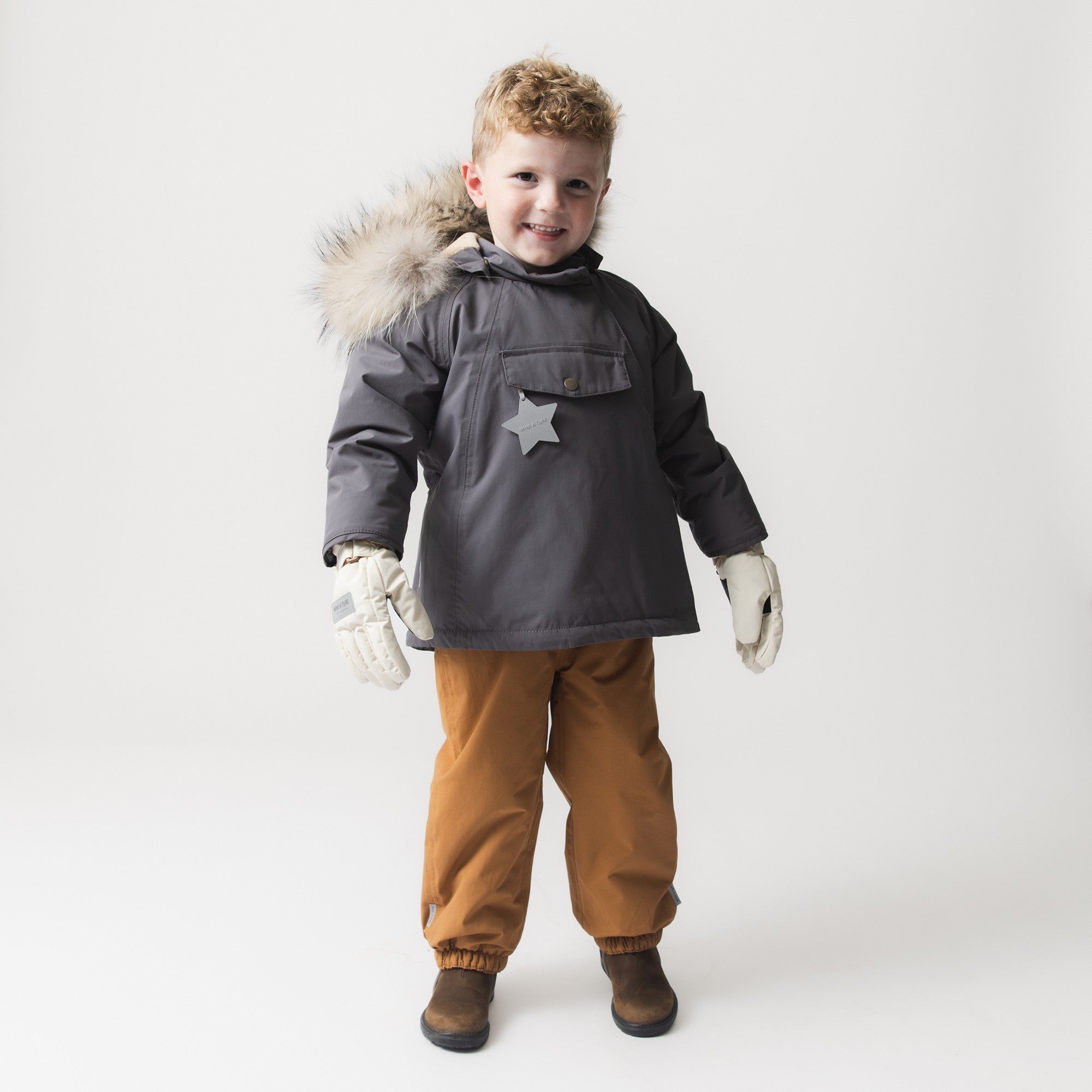 Young child wearing the MATWANG fleece lined winter jacket in charcoal grey with a detachable real fur-trimmed hood, reflective star pocket detail, paired with mustard trousers and cream mittens.