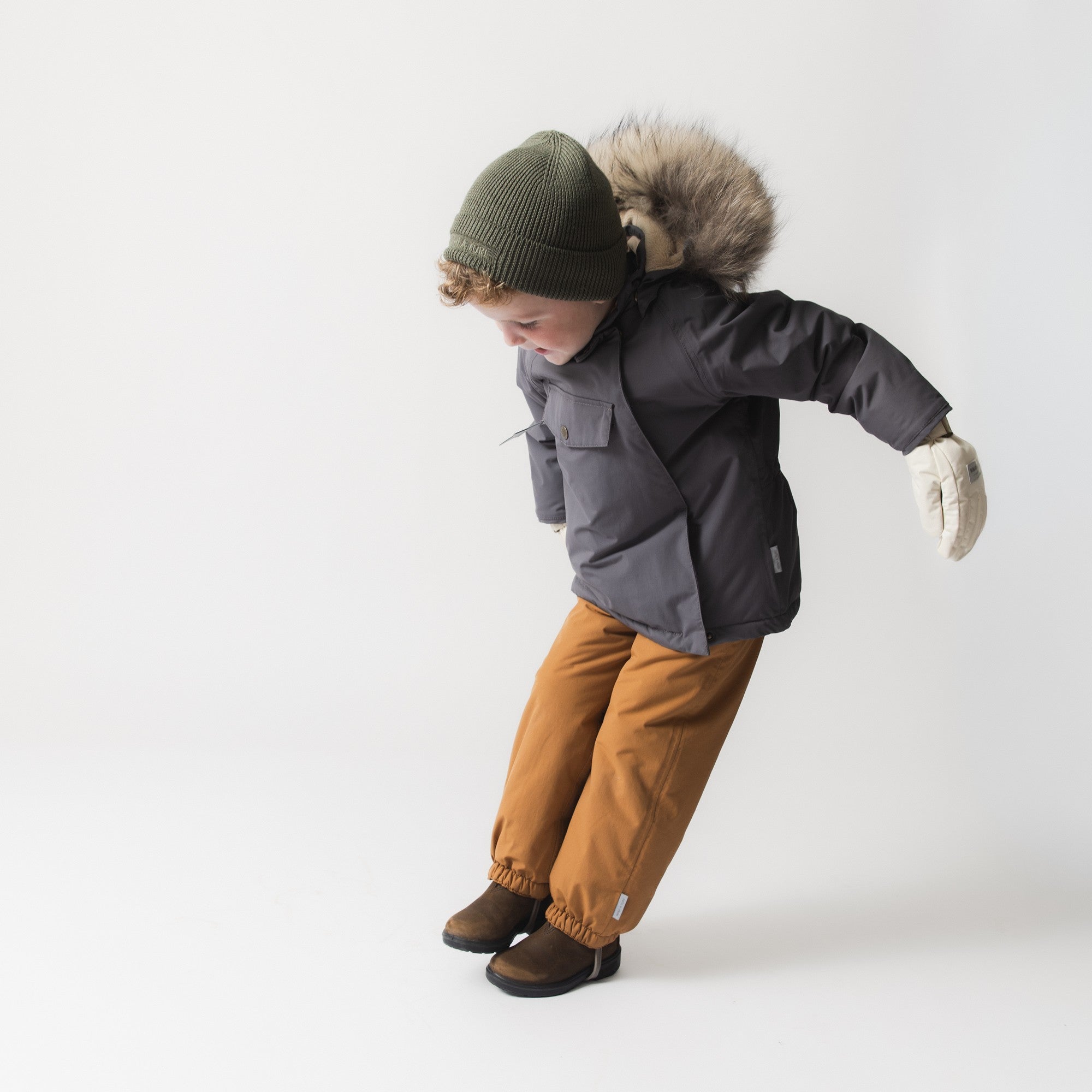 Child in motion wearing the MATWANG fleece lined winter jacket in charcoal and grey with a detachable real fur hood, paired with ochre trousers, cream mittens and a khaki knit beanie, demonstrating the jacket's freedom of movement and improved sleeve fit.
