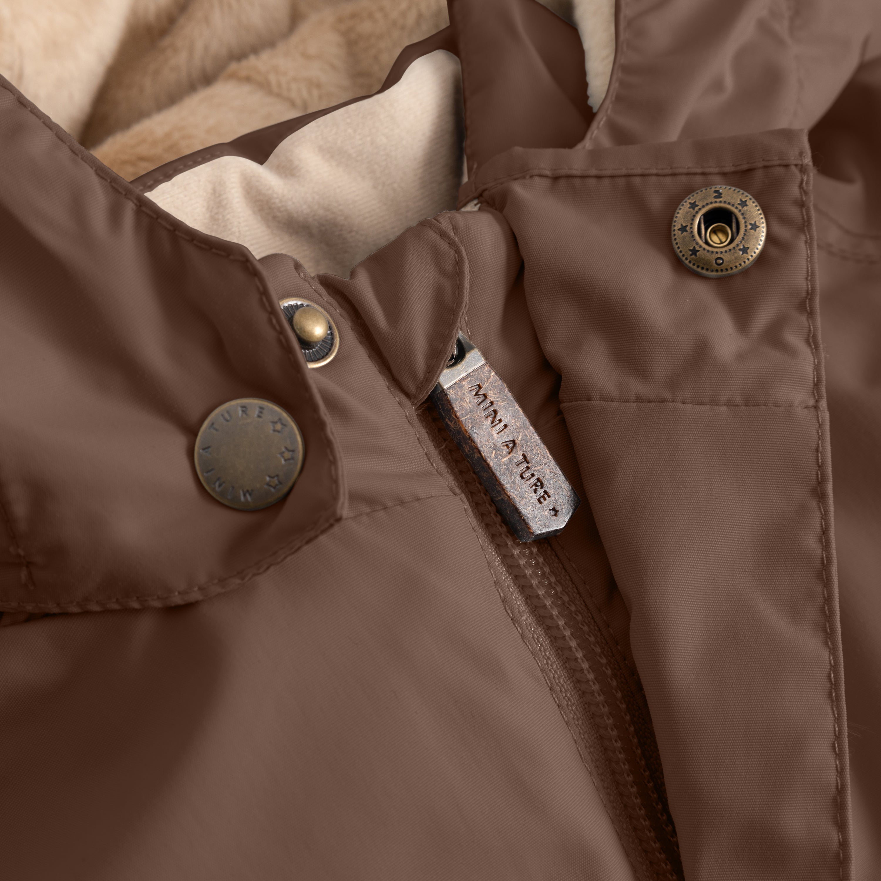 Close-up of the MATWALLY fleece lined winter jacket in brown, showing the hidden centre-front zip with metal MINI A TURE branded zip pull, snap button placket with embossed logo, beige fleece lining visible inside and the angled front pocket with snap button closure.