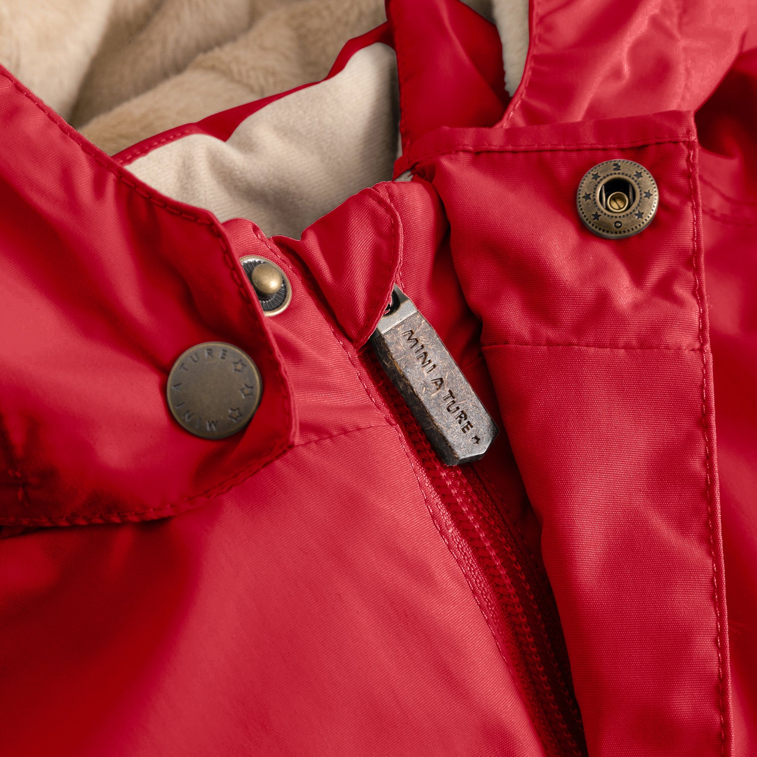 Close-up of the MATWALLY fleece lined winter jacket in red, showing the metal zip pull engraved with MINI A TURE branding, snap button placket with embossed logo detail, beige fleece lining visible inside the collar and the angled front pocket with snap button closure.