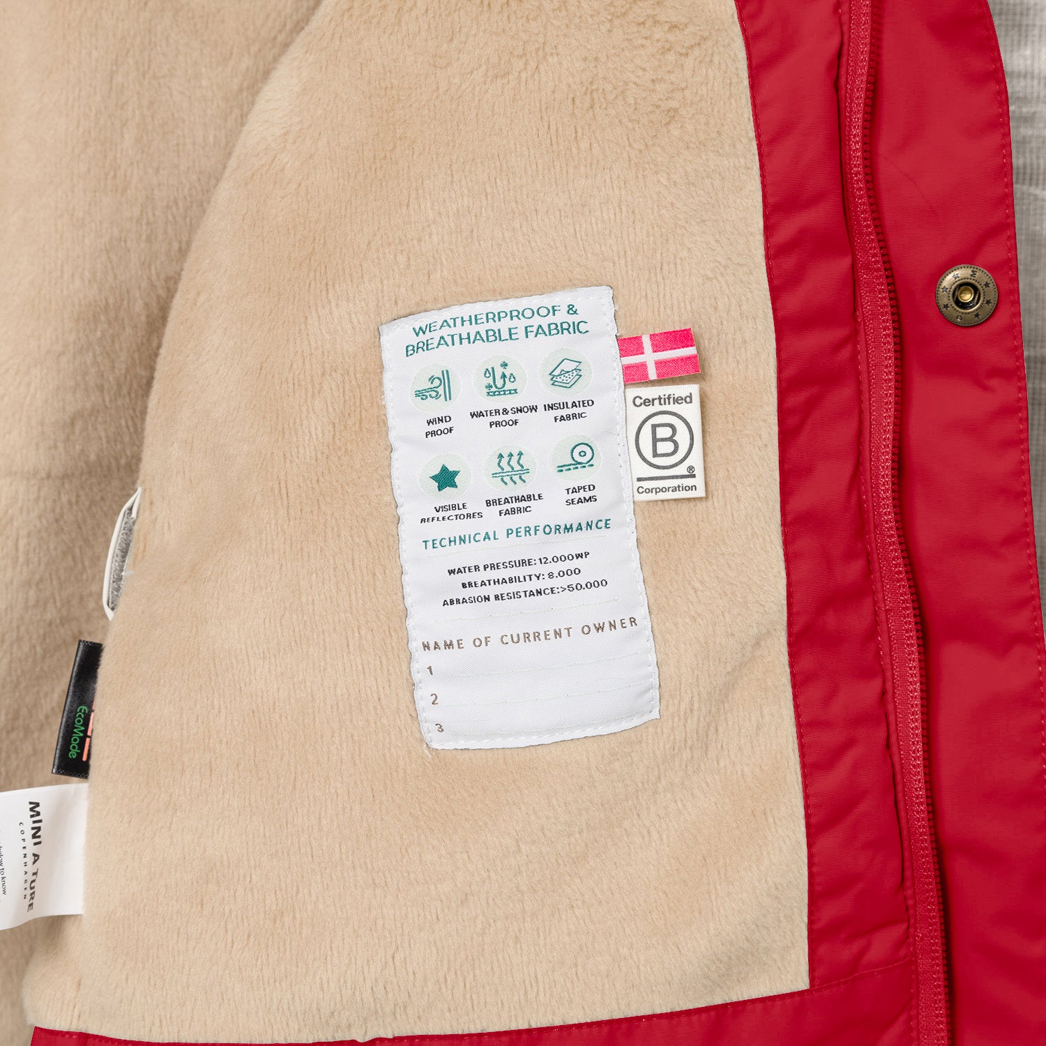 Interior view of the MATWALLY fleece lined winter jacket showing the beige fleece lining with a sewn-in technical performance label displaying waterproof, breathable and insulation icons, Danish flag, B Corp certification logo, metal snap button with embossed branding and a handwritten owner name section at the lower edge.