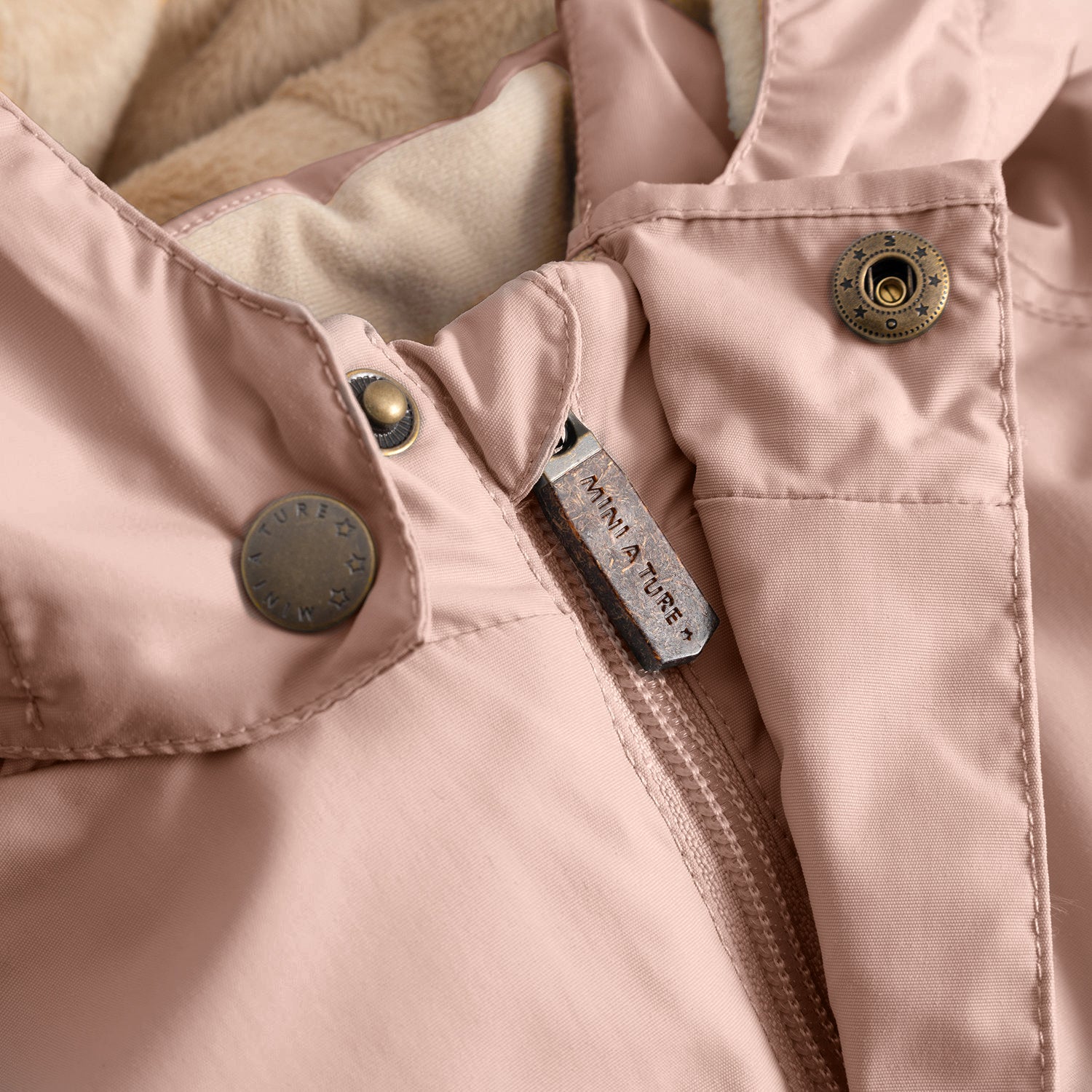 Close-up of the MATWALLY fleece lined winter jacket in dusty pink, showing the metal snap button with embossed MINI A TURE branding, embossed leather zip pull tab with MINI A TURE text, beige fleece lining visible at the collar edge and the fully taped seam construction along the placket.