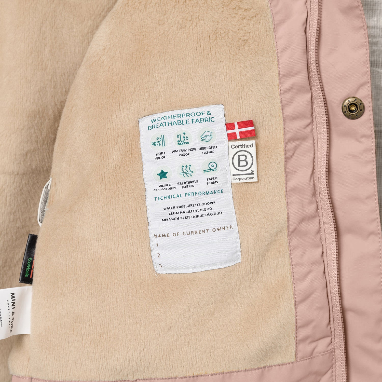 Interior view of the MATWALLY fleece lined winter jacket showing the beige fleece lining with a sewn-in technical performance label displaying waterproof, breathable and insulation specifications, Danish flag, B Corp certification logo and numbered owner name section, alongside the dusty pink shell with metal snap button and concealed zip.