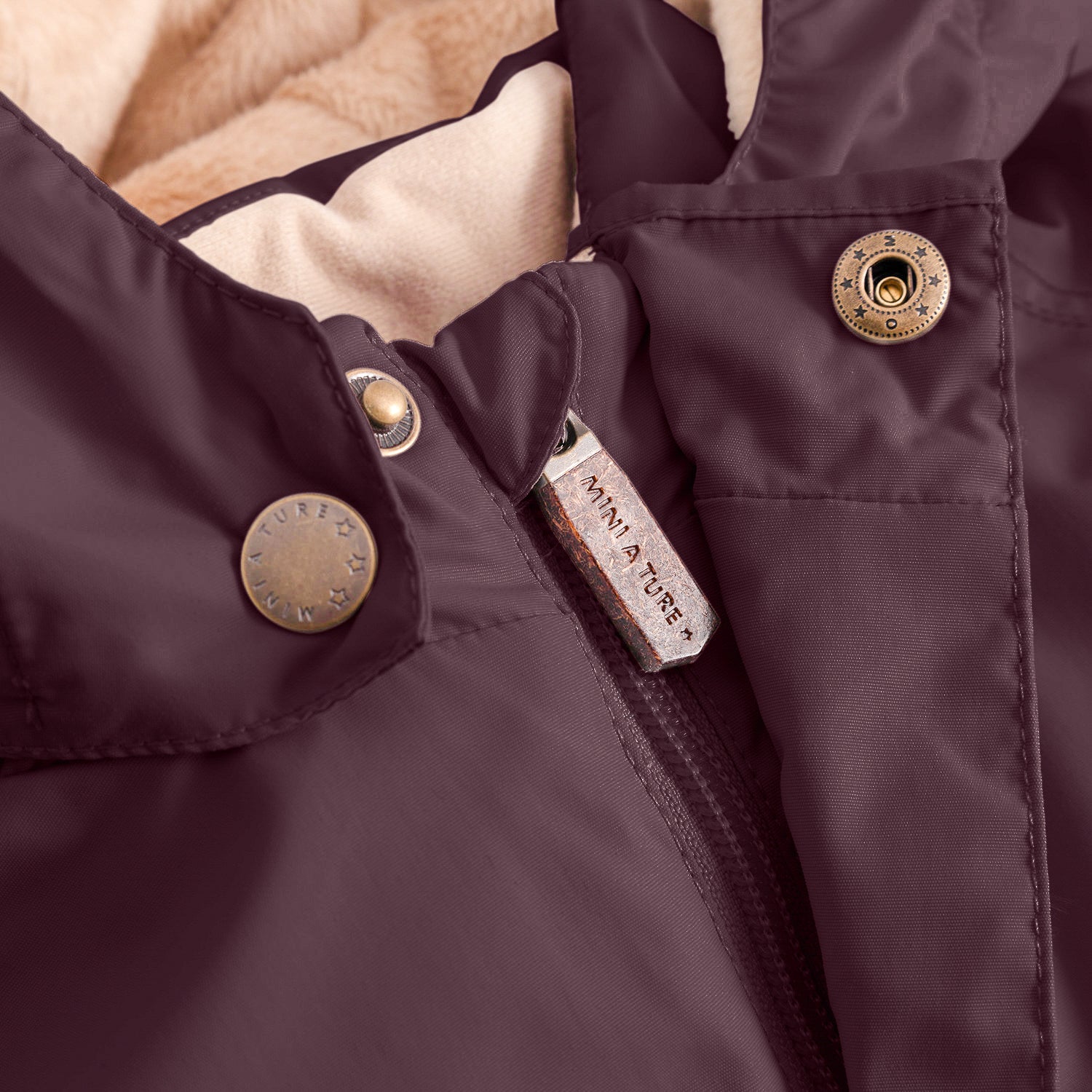 Close-up of the MATWALLY fleece lined winter jacket in deep burgundy, showing the concealed centre-front zip with embossed MINI A TURE leather pull tab, metal snap button with embossed MINI A TURE branding, angled front pocket with snap closure and beige fleece lining visible at the collar edge.