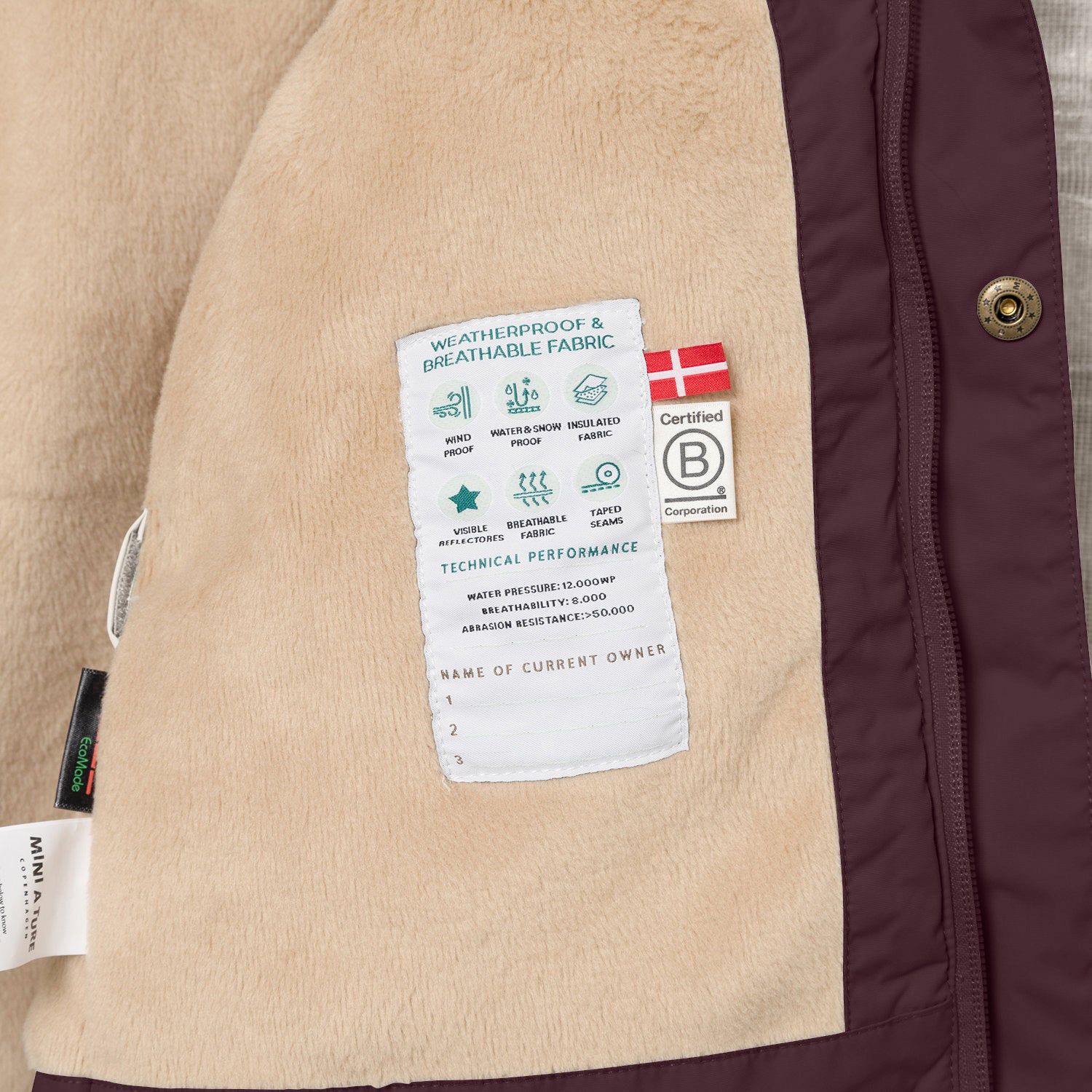 Interior detail of the MATWALLY fleece lined winter jacket showing the beige fleece lining with a sewn-in technical performance label displaying waterproof, breathable and insulation specifications, Danish flag, B Corp certification logo and numbered owner name section, positioned next to the deep burgundy shell with metal snap button and concealed zip.