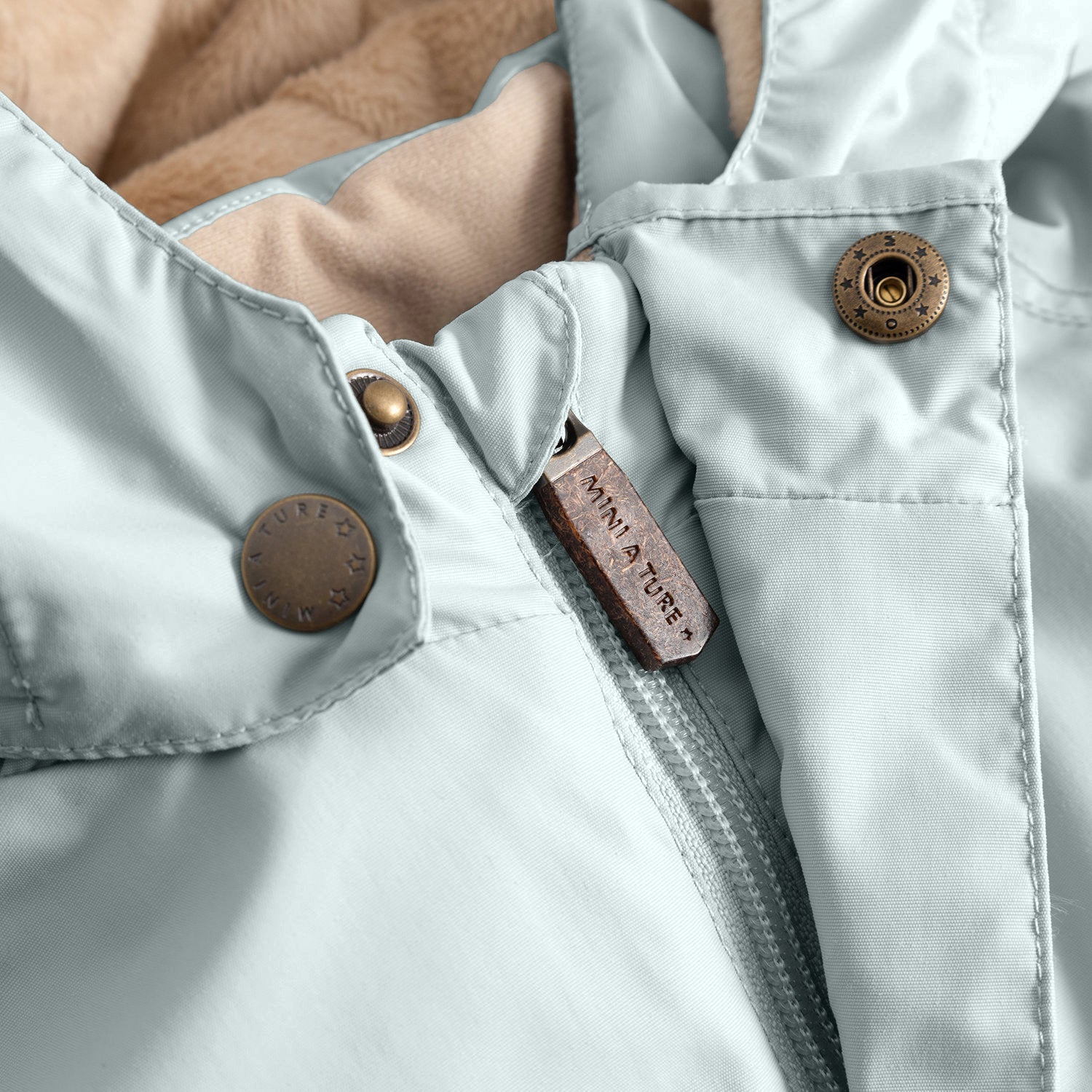 Close-up of the MATWALLY fleece lined winter jacket in pale mint green, showing the concealed centre-front zip with embossed MINI A TURE leather pull tab, metal snap buttons with embossed MINI A TURE branding and beige fleece lining visible at the collar edge.