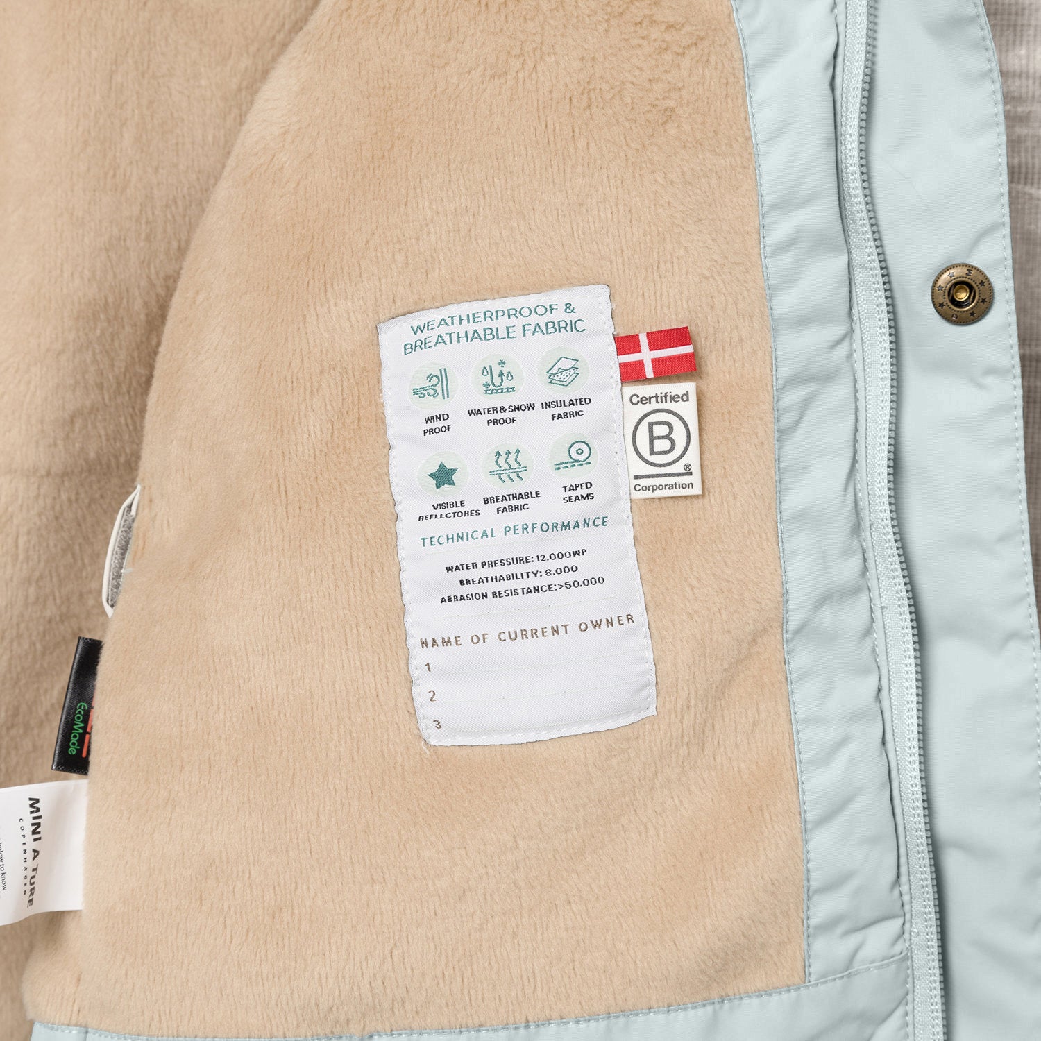 Interior detail of the MATWALLY fleece lined winter jacket showing the beige fleece lining with a sewn-in technical performance label displaying waterproof, breathable and insulation specifications, Danish flag, B Corp certification logo and numbered owner name section, positioned next to the pale mint green shell with metal snap button and concealed zip.