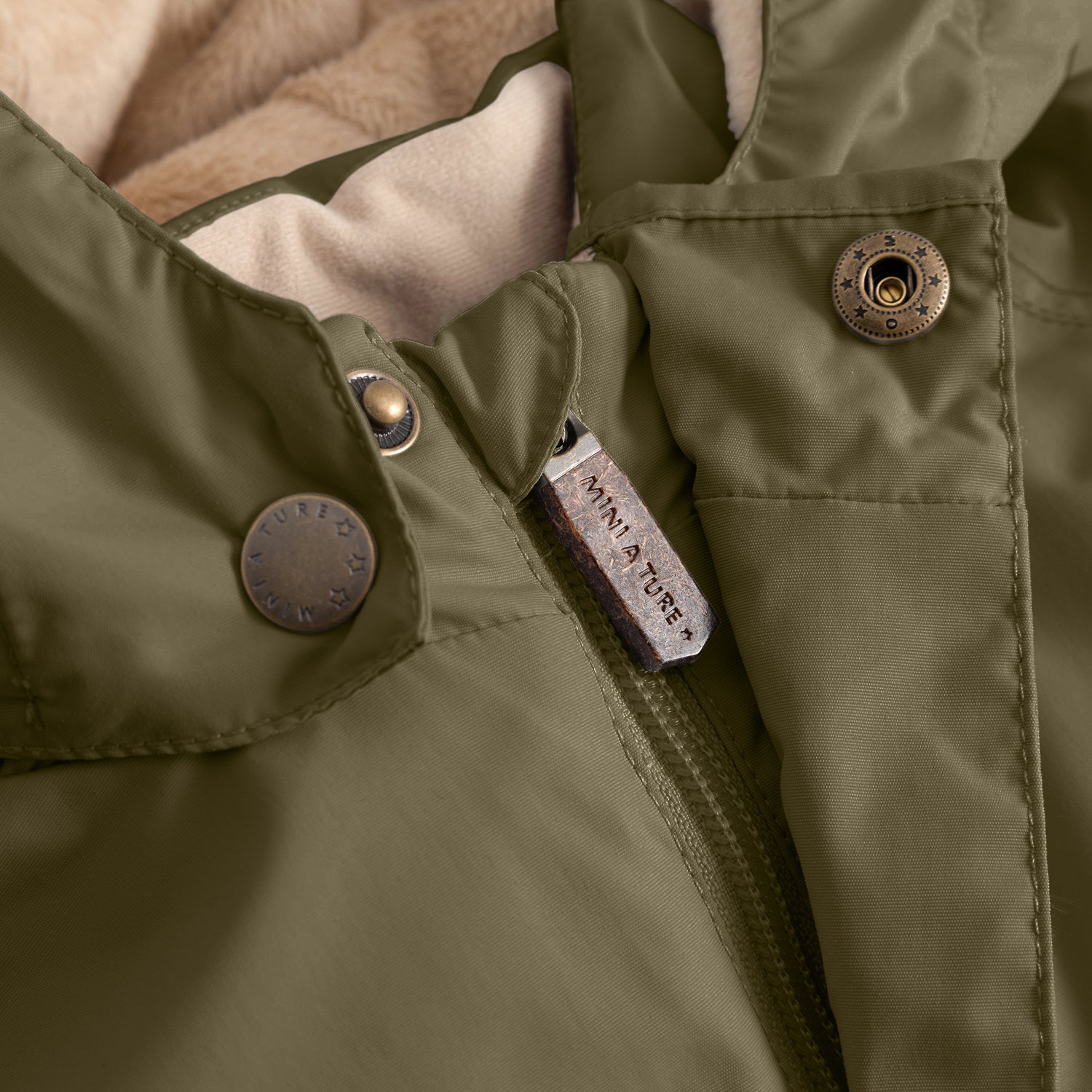 Detail of the MATWALLY fleece lined winter jacket showing the MINI A TURE branded zip pull, snap button fastening on the detachable hood and the beige fleece lining visible at the collar.