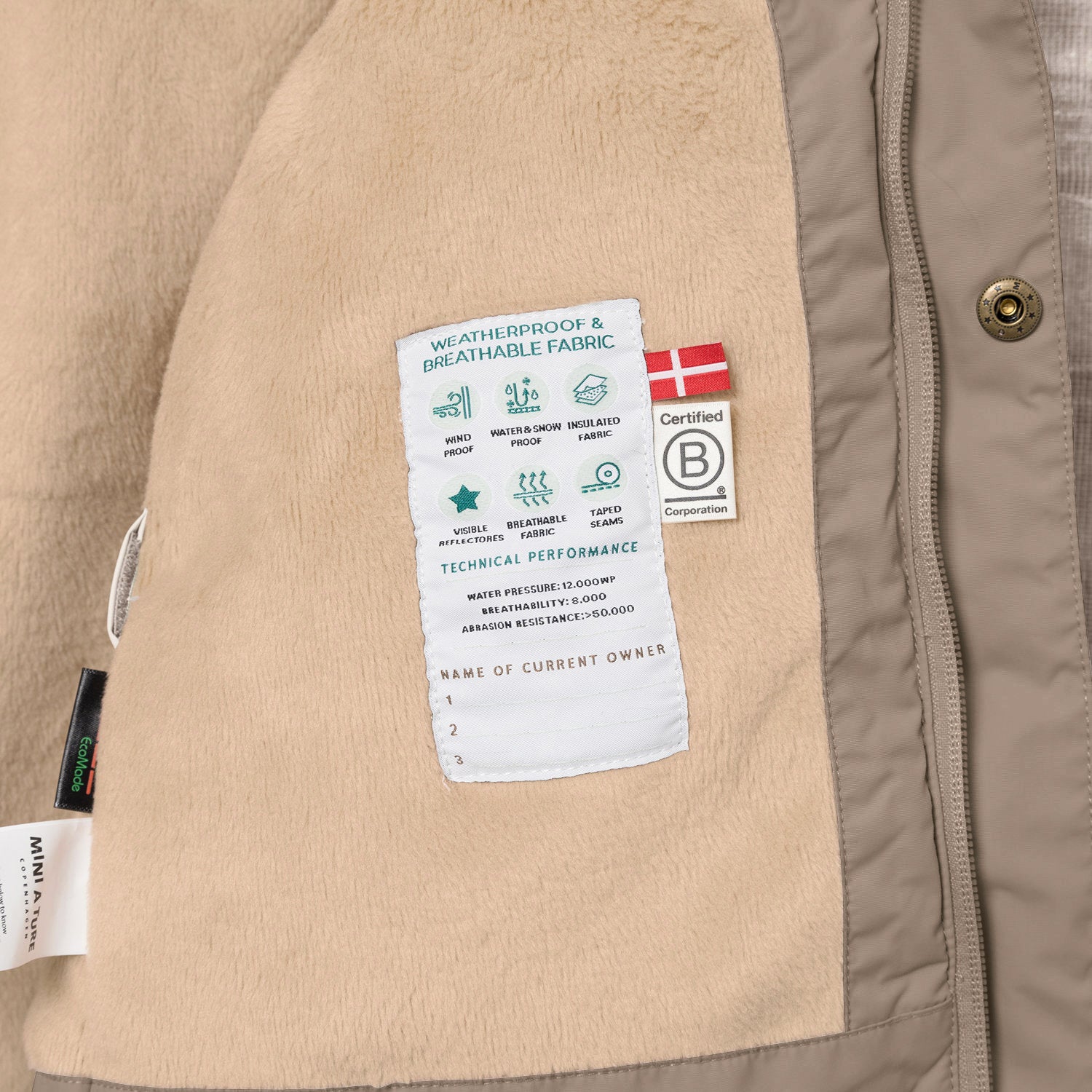 Interior view of the MATWALLY fleece lined winter jacket in taupe, showing the beige fleece lining with a sewn-in technical performance label displaying windproof, waterproof and breathable icons, Danish flag, B Corp certification logo, and a handwritten owner name section.