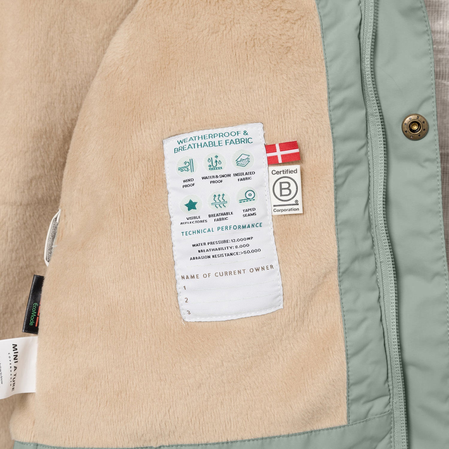 Interior view of the MATWALLY fleece lined winter jacket showing the beige fleece lining with a sewn-in technical performance label displaying waterproof, breathable and insulation icons, Danish flag, B Corp certification logo and a handwritten owner name section at the lower edge, alongside the sage green shell with metal snap button and hidden zip.