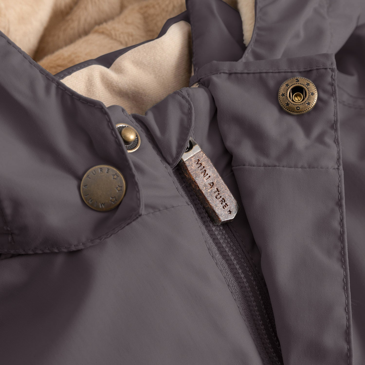 Close-up of the MATWALLY fleece lined winter jacket in charcoal grey, showing the concealed centre-front zip with embossed MINI A TURE metal pull tab, embossed metal snap button securing the angled front pocket, and the beige fleece lining visible at the collar.