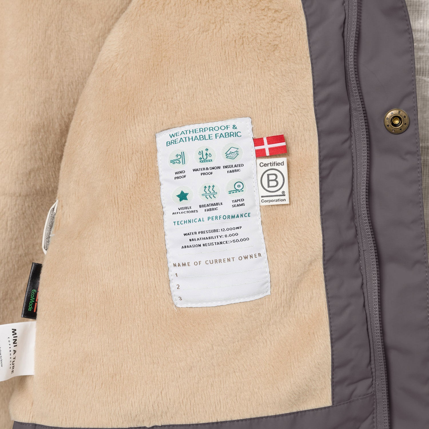 Interior view of the MATWALLY fleece lined winter jacket in charcoal grey, showing the beige fleece lining with a sewn-in white technical performance label displaying waterproof and breathability specifications, Danish flag, B Corp certification logo and numbered owner name section, positioned beside the concealed centre-front zip with embossed metal snap button.
