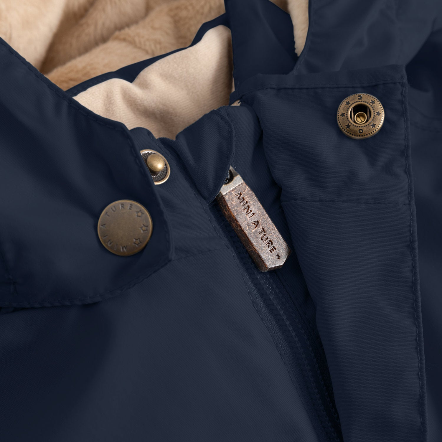 Close-up of the MATWALLY fleece lined winter jacket in navy blue, showing the embossed MINI A TURE metal snap button securing the detachable hood, the concealed centre-front zip with embossed MINI A TURE leather pull tab, and the beige fleece lining visible at the collar.