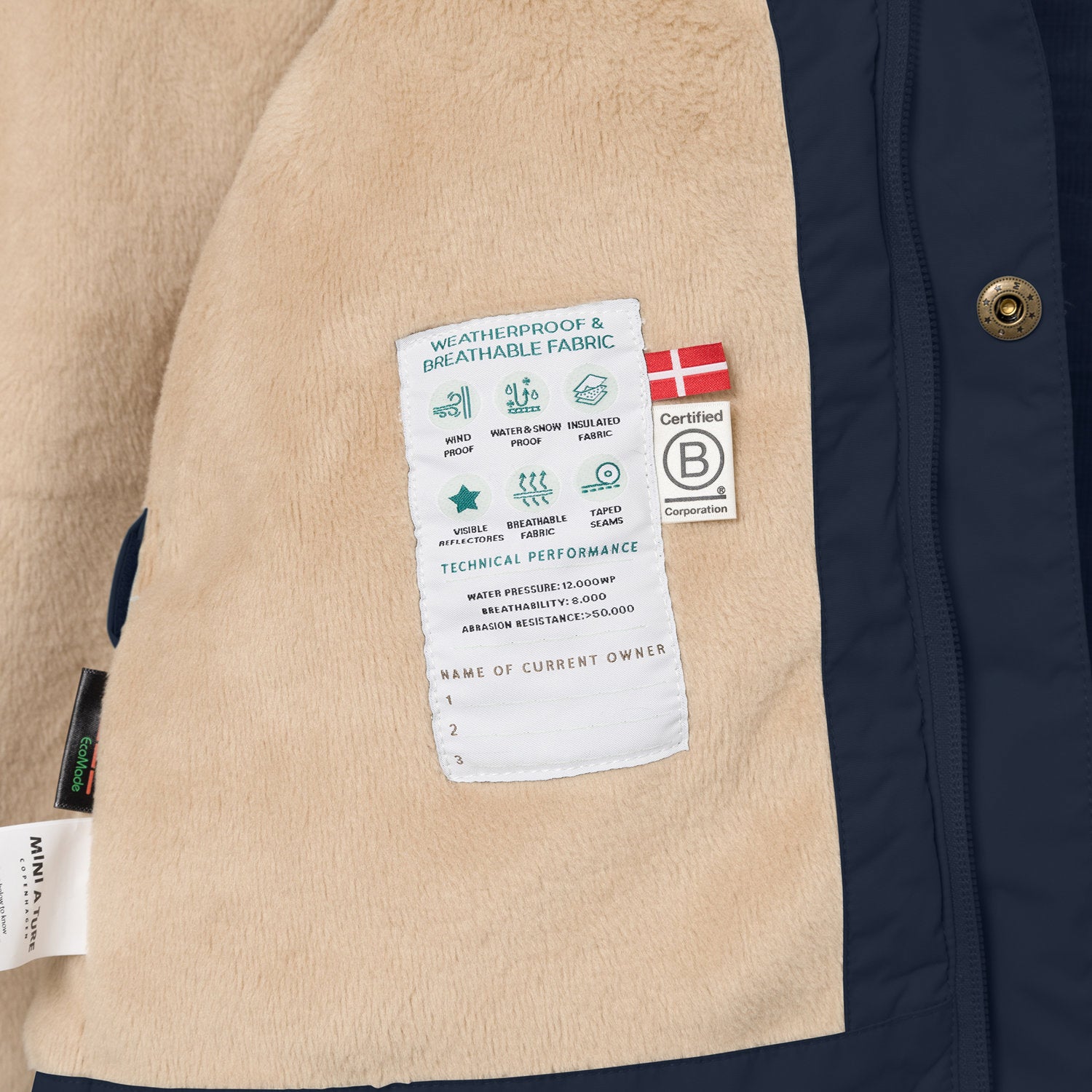 Interior detail of the MATWALLY fleece lined winter jacket showing the beige fleece lining with a sewn-in white technical performance label displaying waterproof and breathability specifications, Danish flag, B Corp certification logo and numbered owner name section, positioned beside the navy blue outer shell with embossed metal snap button.