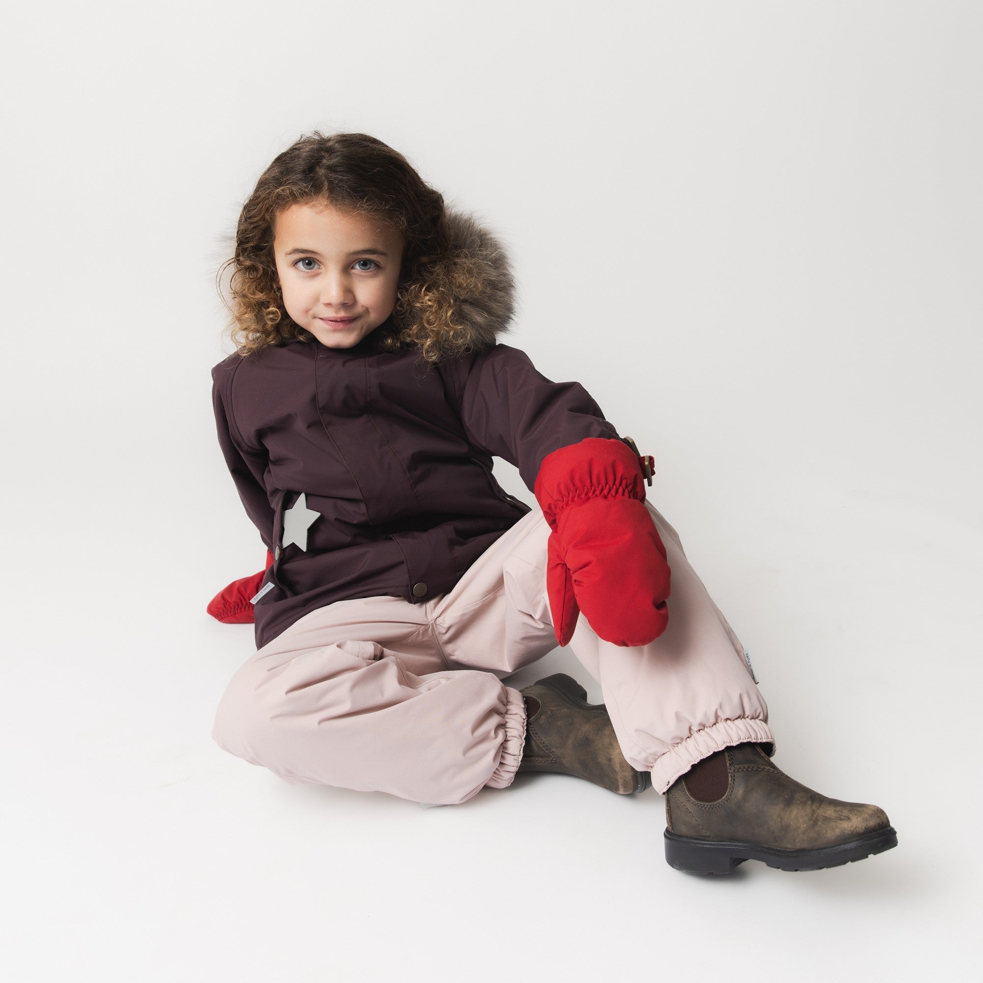 Young child seated wearing the MATWALLY fleece lined winter jacket in burgundy with detachable real fur hood trim, paired with pale pink snow trousers and bright red mittens, showing the jacket's comfortable fit and practical layering for winter play.