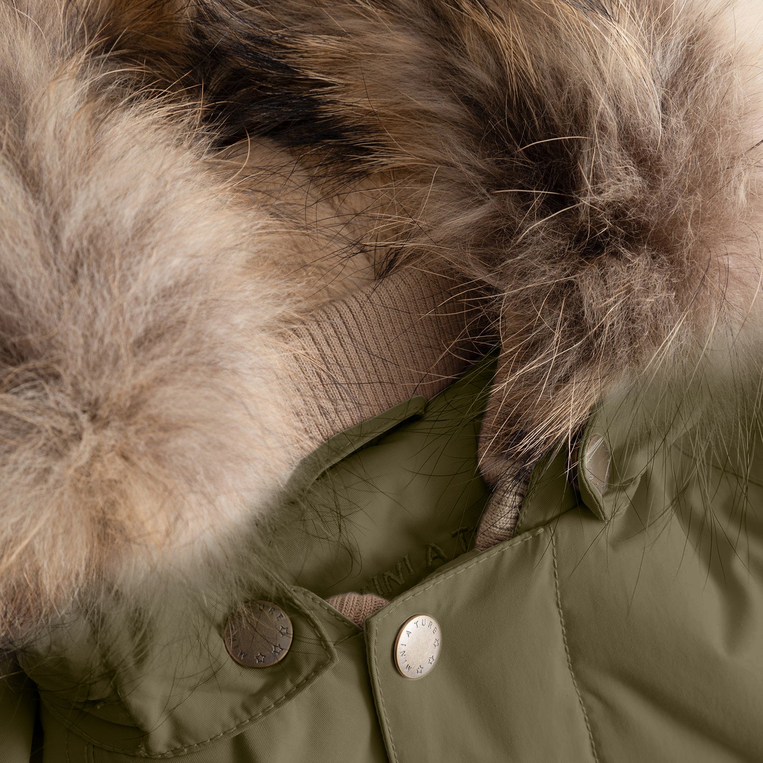 Close-up detail of the MATWALLY fleece lined winter jacket's collar in olive green, highlighting the natural multi-tonal real fur trim with soft, layered texture, branded MINI A TURE press studs securing the detachable hood, and the beige ribbed collar visible beneath the fur.