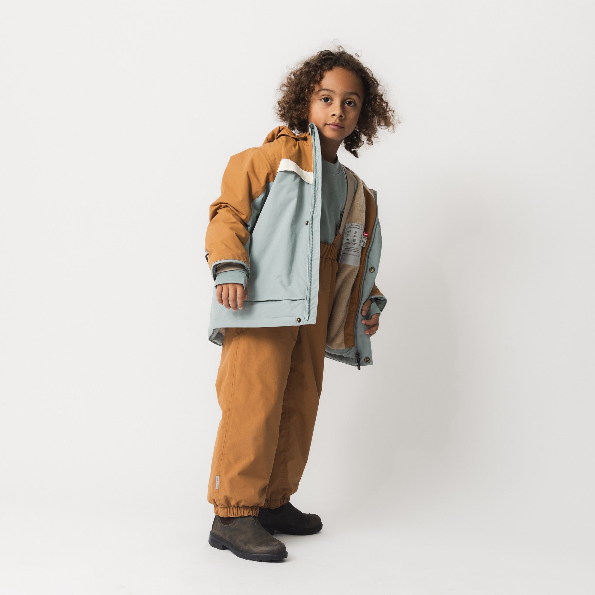 Child holding the MATWALDO fleece lined winter jacket open to display the soft fleece lining, THERMOLITE insulation and adjustable internal waist, while wearing matching waterproof trousers in brown sugar with a reflective detail visible at the lower leg.