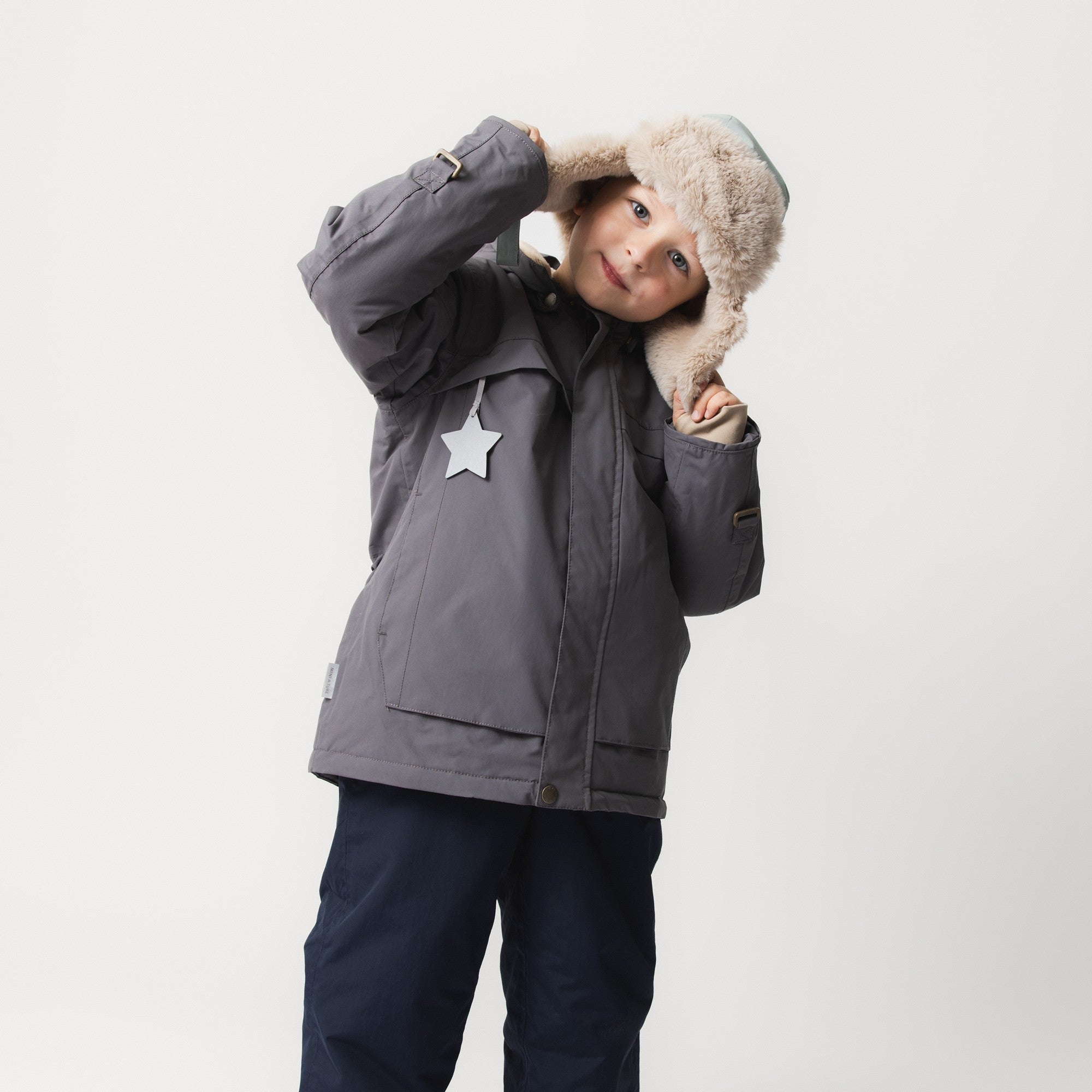 MATWALDO fleece lined winter jacket in charcoal grey shown from the back with the detachable hood fastened, metal D-ring details on both sleeves, beige fleece lining visible at the cuffs and a white star-shaped reflective detail attached to the left sleeve.